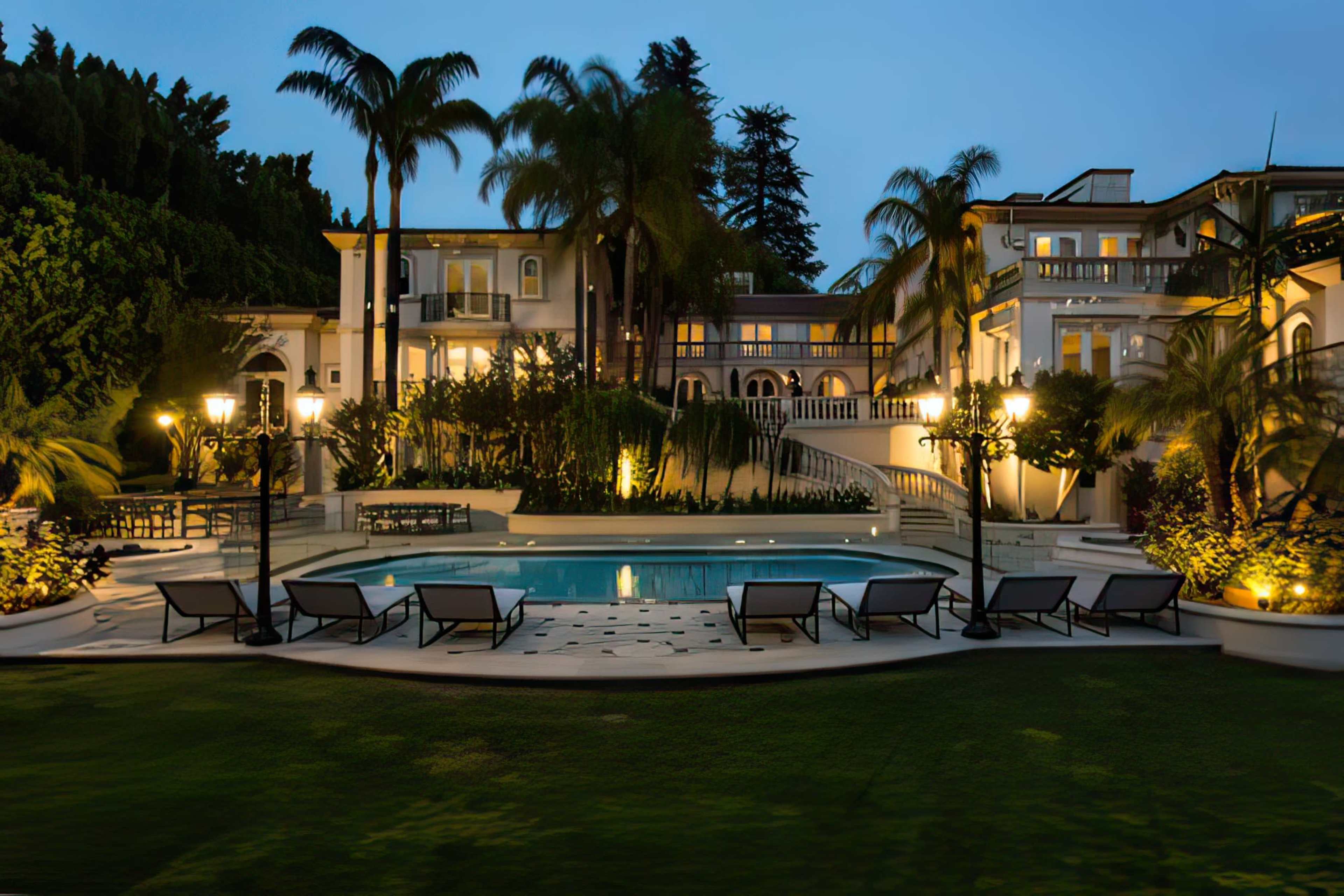 A spacious mansion with a swimming pool is surrounded by palm trees and illuminated by outdoor lighting at dusk.