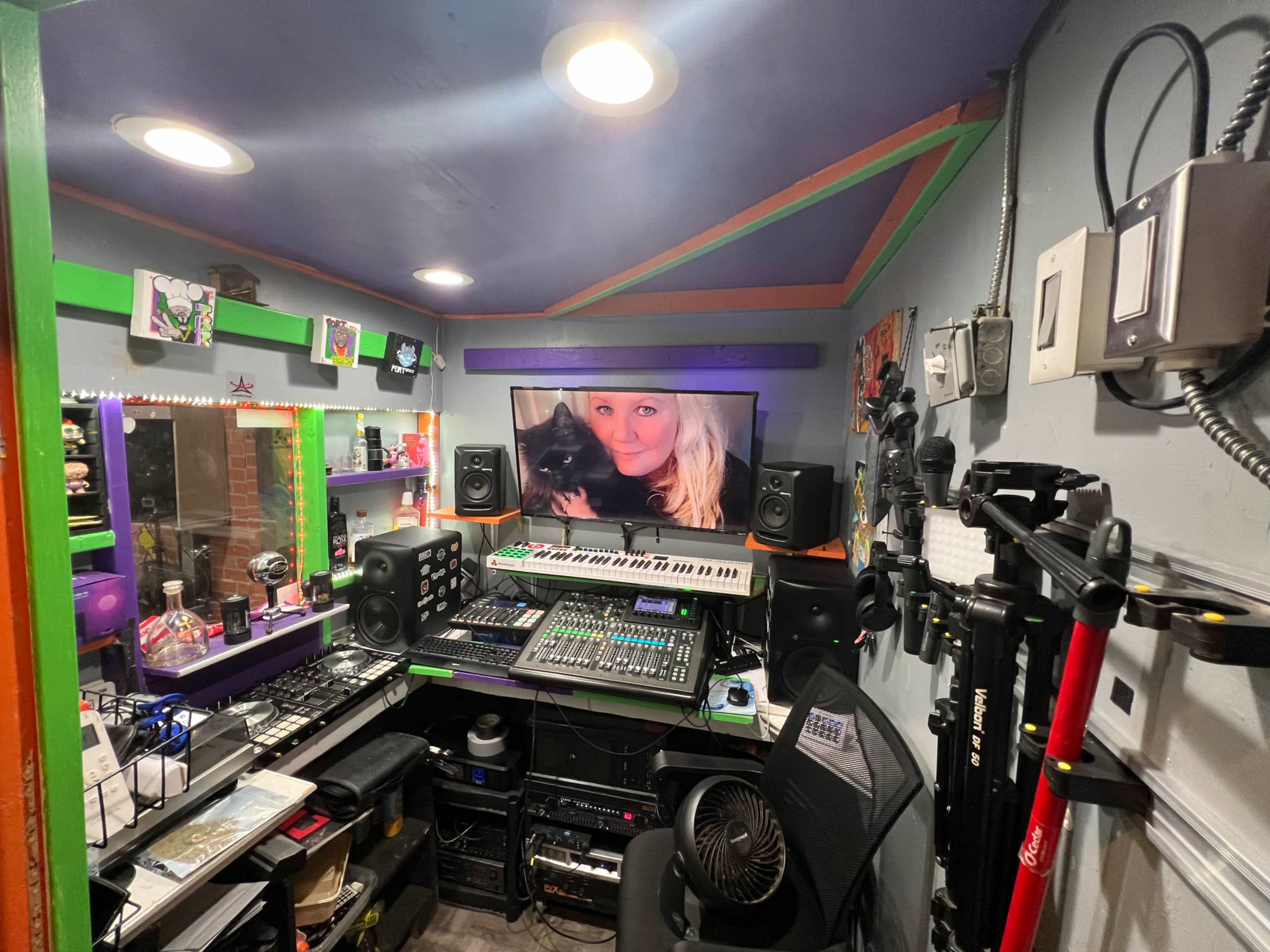 A small recording studio features a digital audio workstation setup with sound equipment, a large screen displaying a woman with a cat, and colorful walls.
