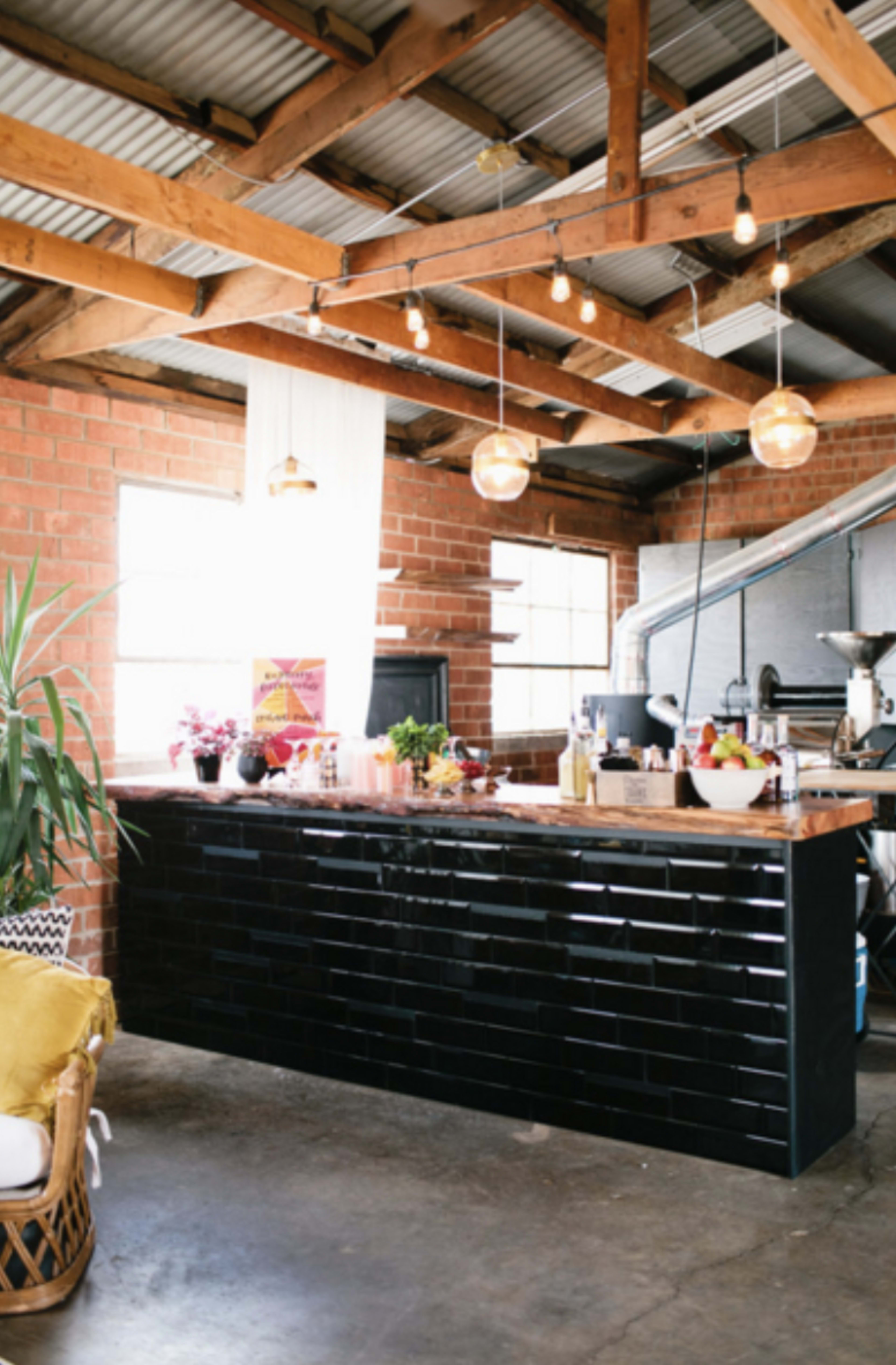 The image features a modern kitchen space with a black tiled island, wooden beams overhead, and pendant lighting, set against a backdrop of exposed brick and large windows.