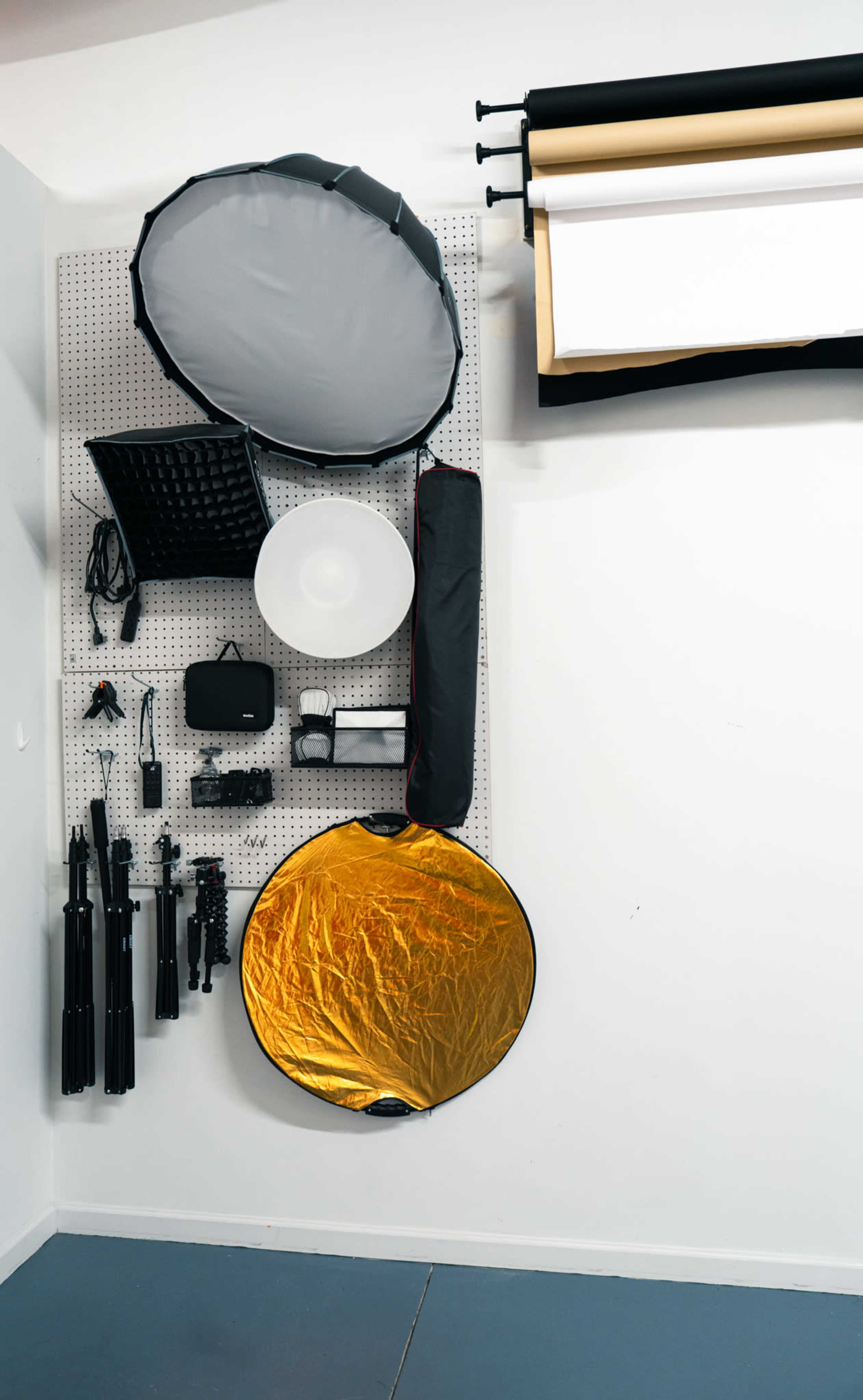 The image shows a wall-mounted pegboard displaying various photography equipment, including lighting modifiers, tripods, and backdrop rolls.