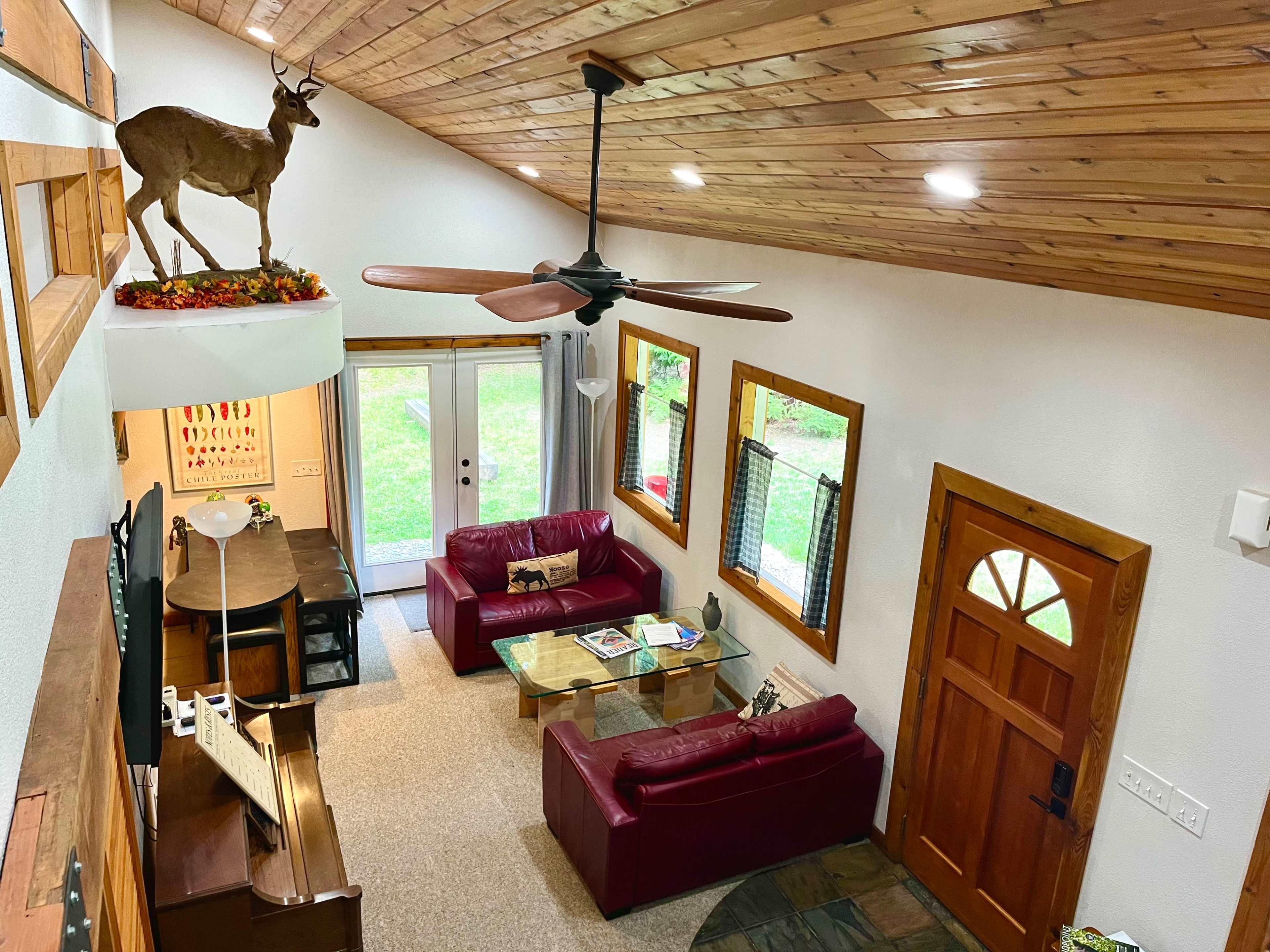 A cozy living room with red couches, a wooden dining table, and a mounted deer head decoratively placed above a door, all situated beneath a vaulted wooden ceiling.