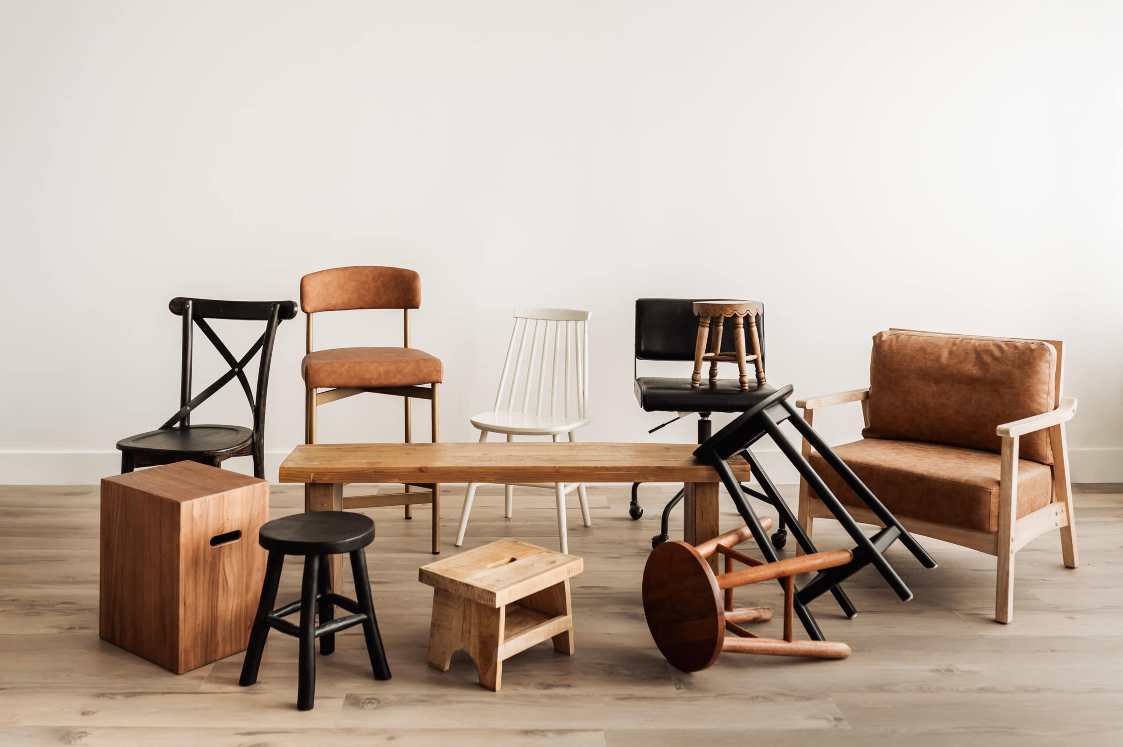The image shows a variety of chairs and stools arranged around a wooden bench on a light-colored wooden floor against a plain white wall.
