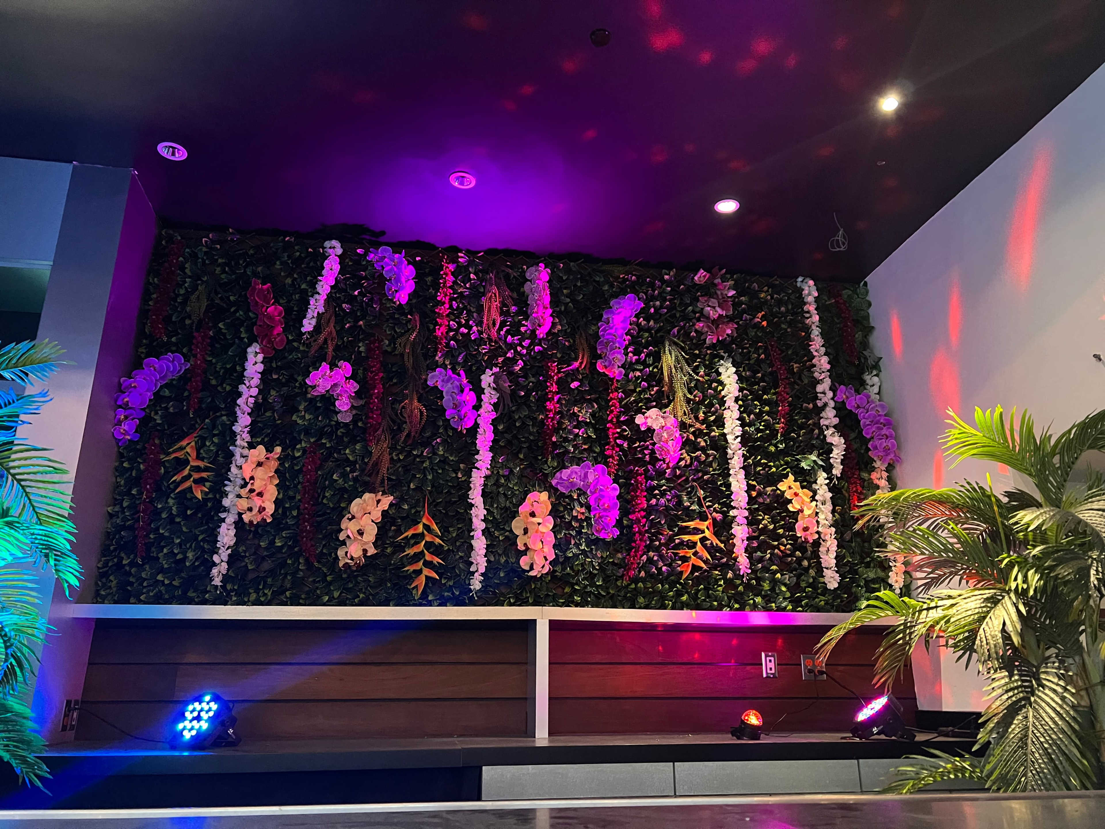 A decorative wall adorned with various artificial flowers and greenery, illuminated by colored lights.