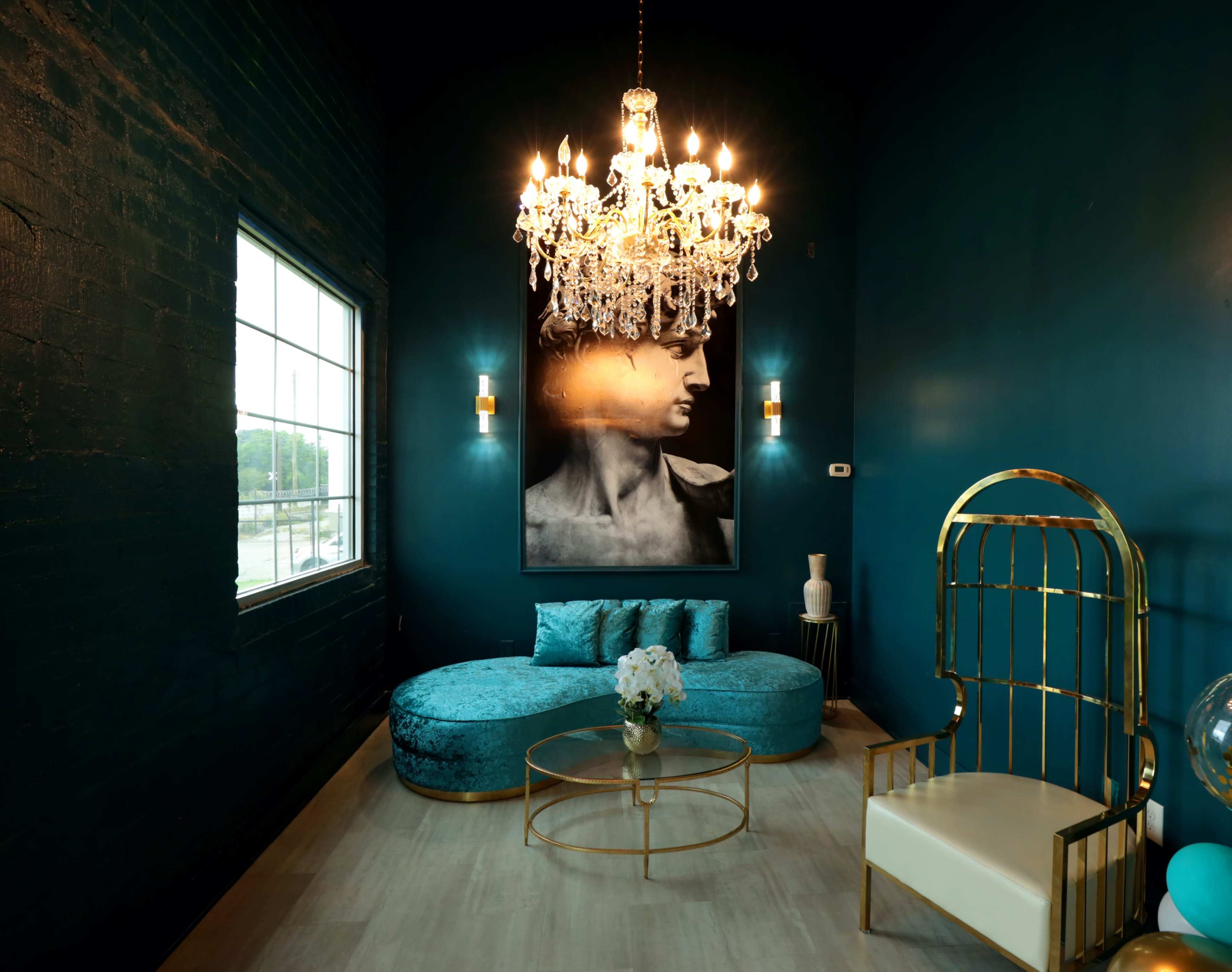 A stylish lounge area with a large chandelier, a teal-colored curved sofa, a golden birdcage chair, and a prominent black-and-white artwork of a statue on the wall.