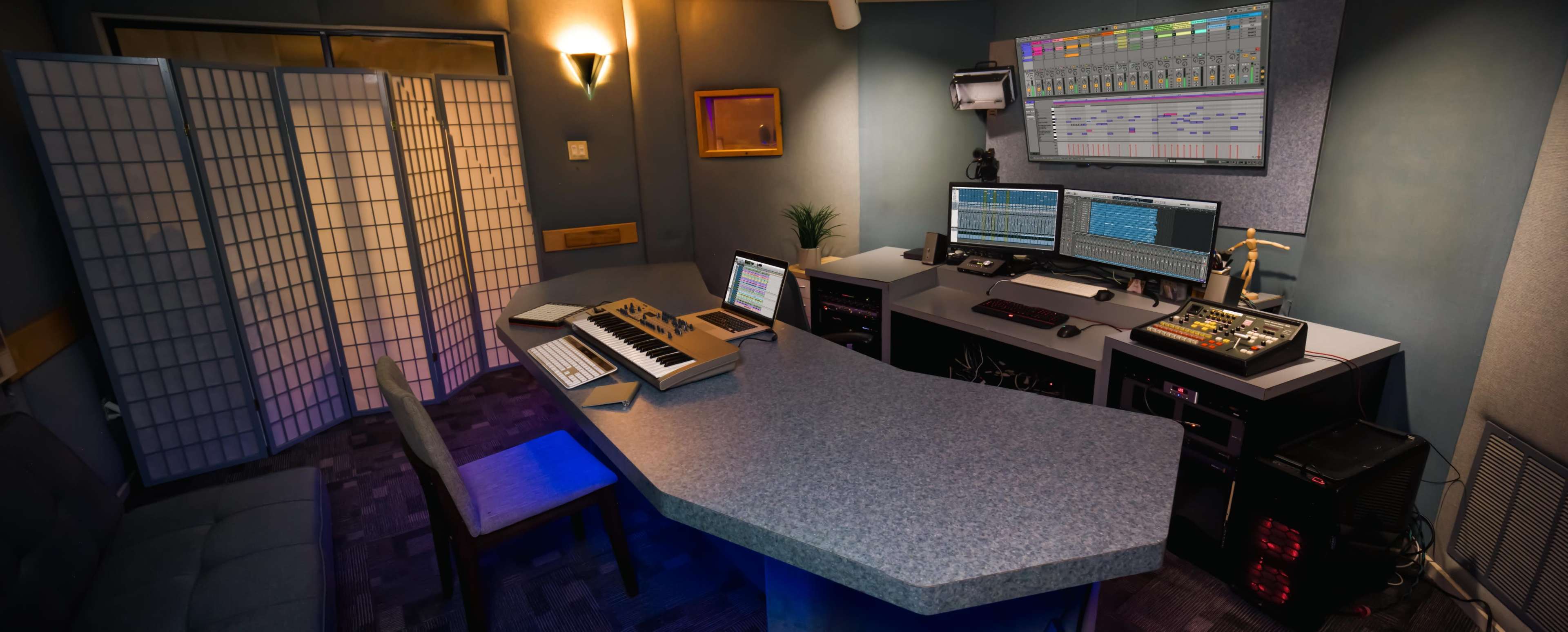 A modern music studio with a central workstation, multiple computer monitors, a keyboard, and sound equipment set against a backdrop of blue-gray walls and decorative screens.