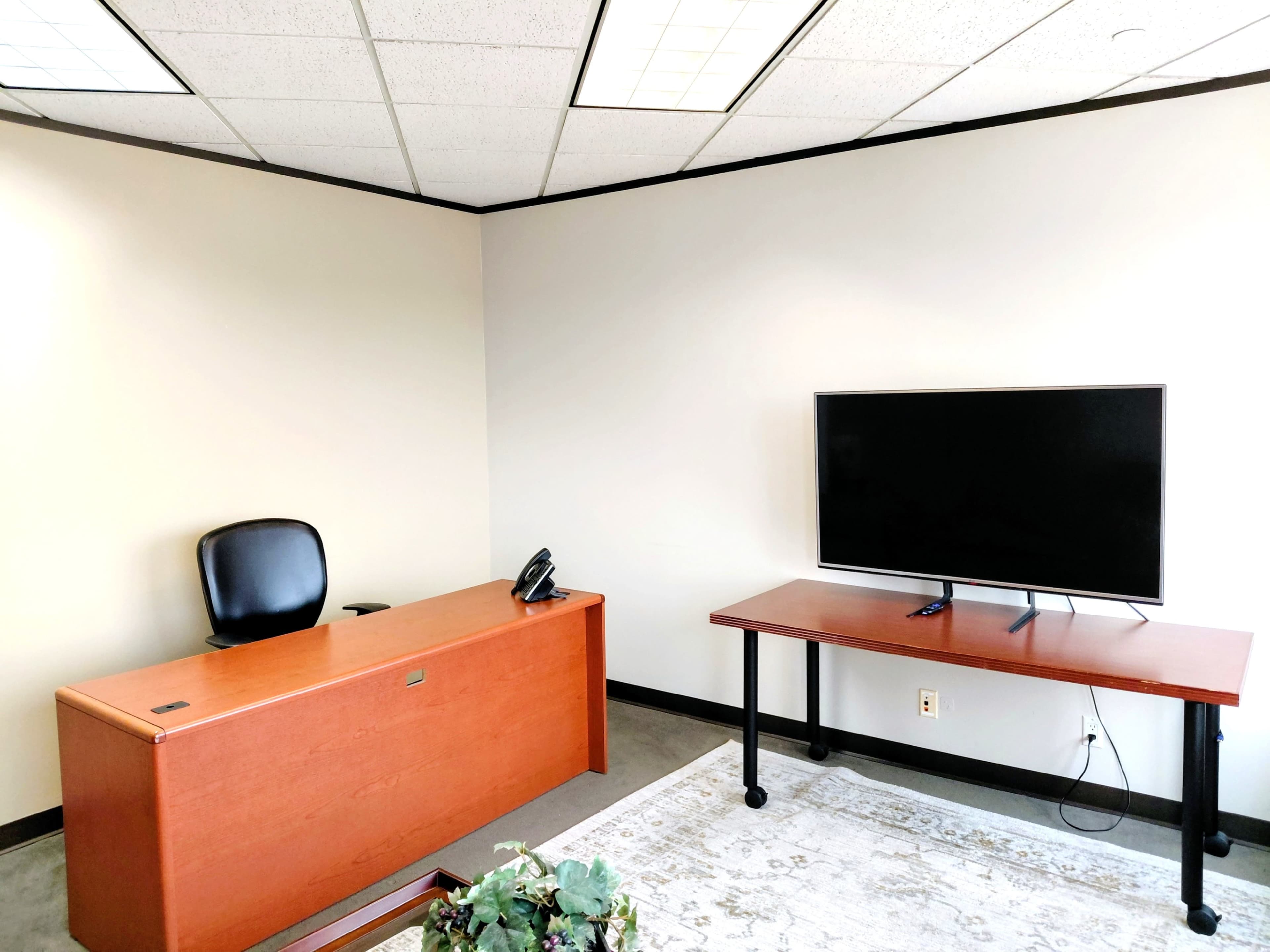 The image shows a minimalistic office space with a wooden desk, a black office chair, a phone, and a television mounted on a separate table.