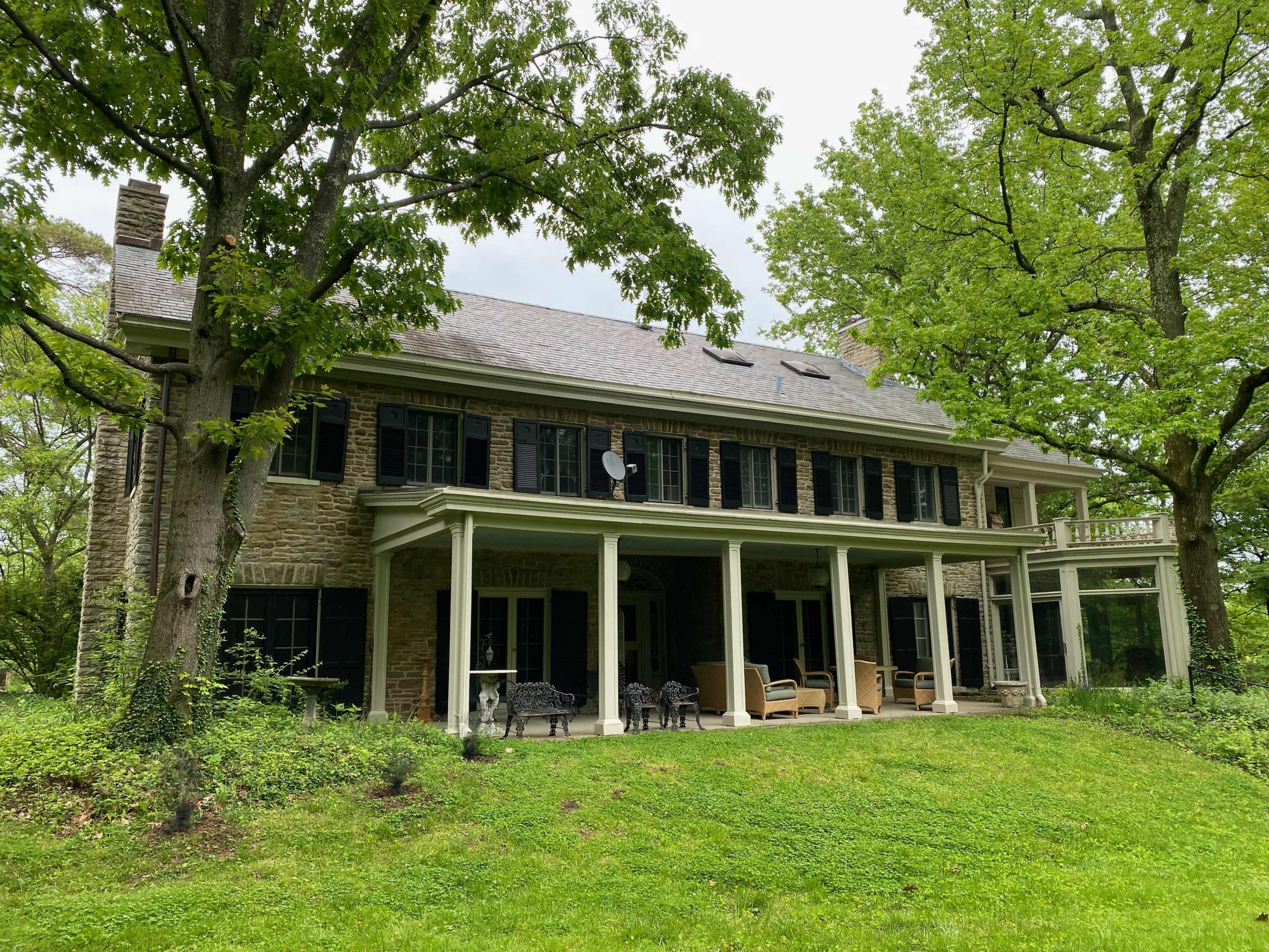 A large two-story stone house with dark shutters and a covered porch, set among green trees and grass.