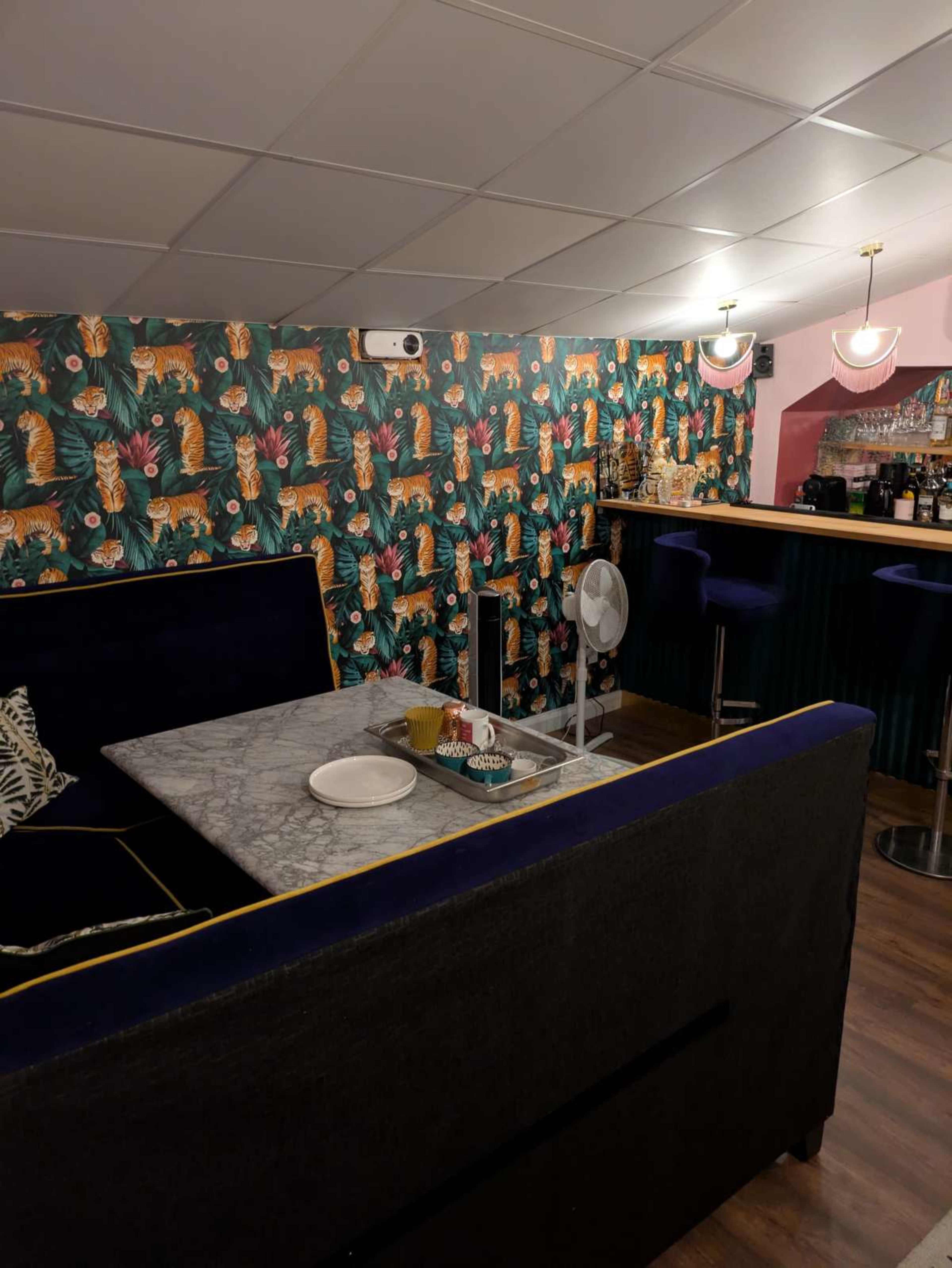 The image shows a cozy room featuring a dark sofa with a marble coffee table, a vibrant tiger-patterned wallpaper, and a bar area with illuminated shelves.