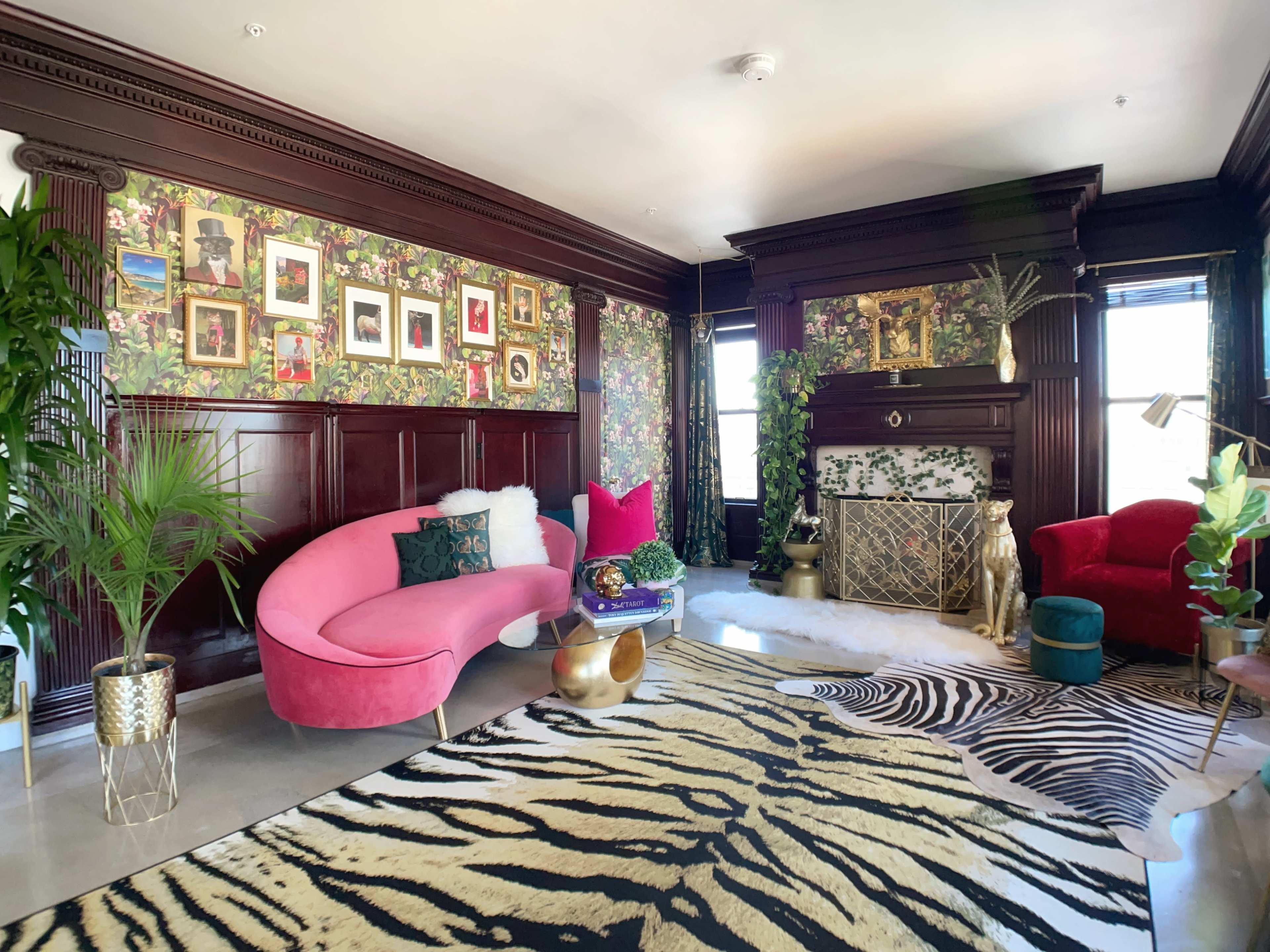 The room features a pink curved sofa, a red armchair, and a zebra-patterned rug, with walls adorned by floral wallpaper and framed pictures.