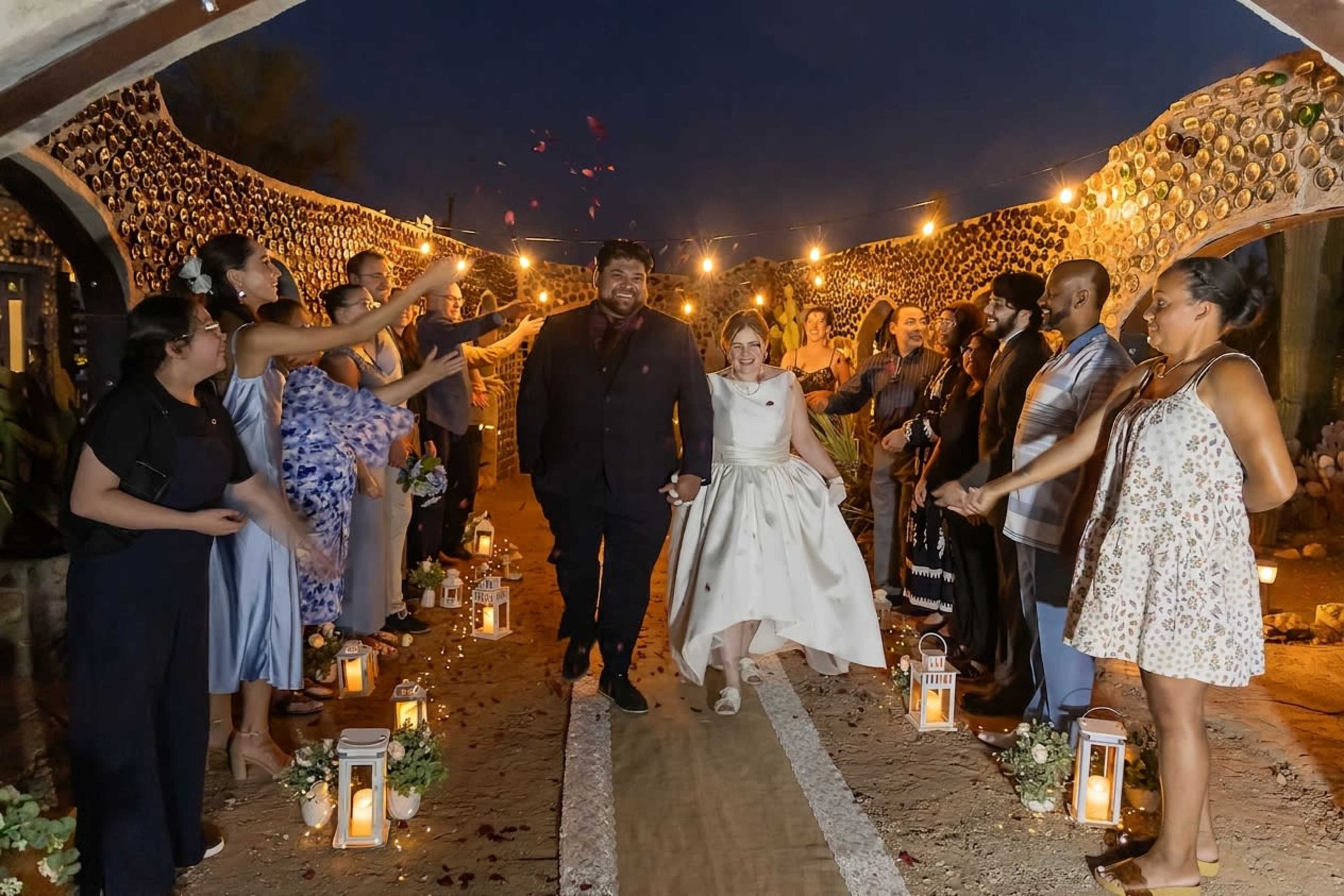 A couple walks down an outdoor aisle lined with candles and flower petals, surrounded by guests celebrating at night.