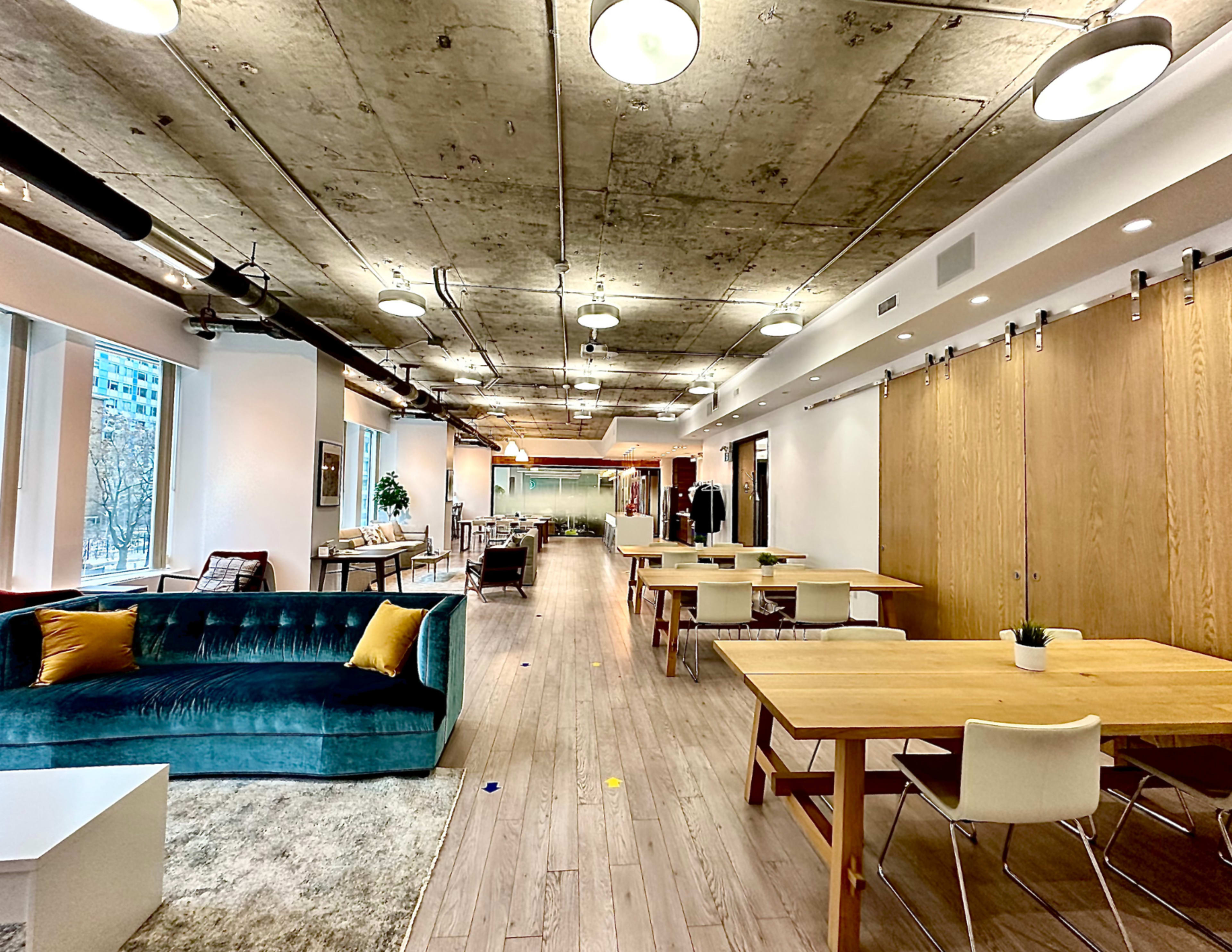 A modern office space with a long hallway featuring wooden tables, a blue couch, and large windows.