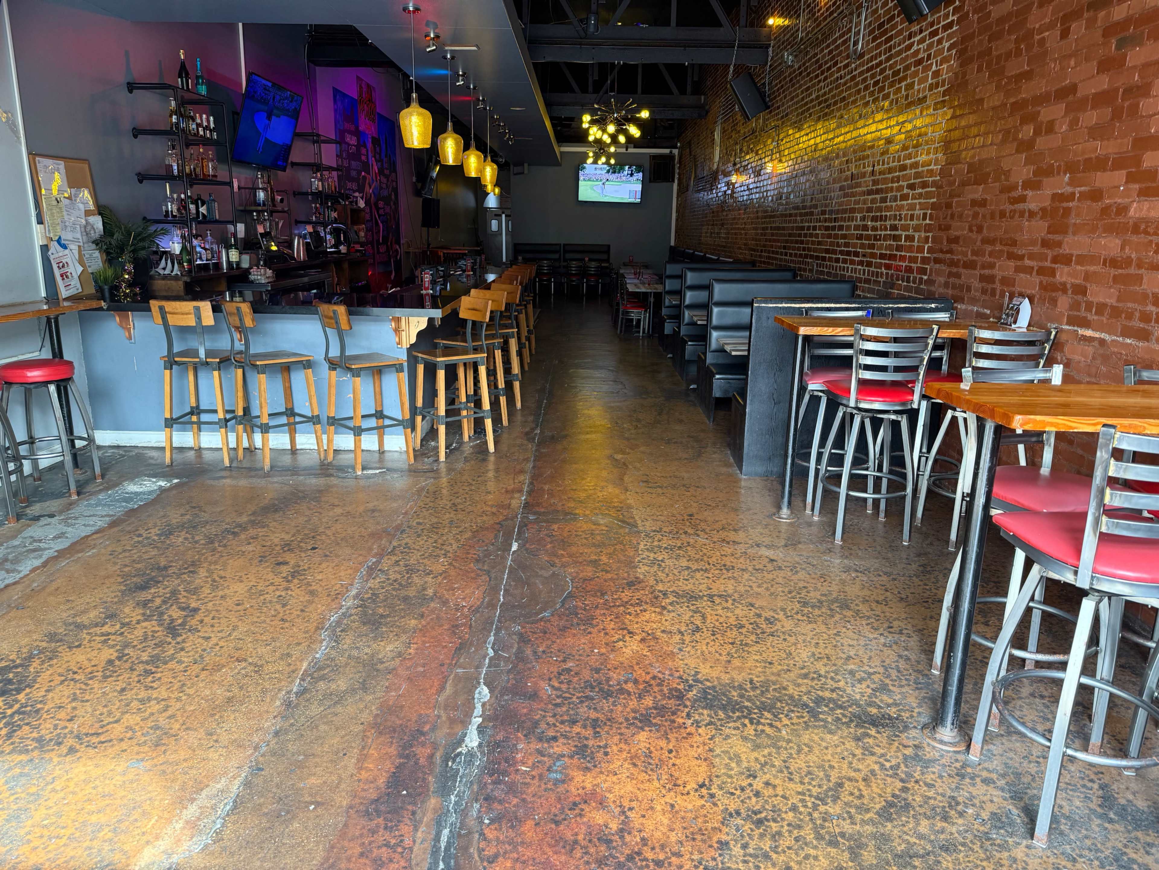 The image shows an empty restaurant interior with wooden tables, metal chairs, a bar area, and exposed brick walls.