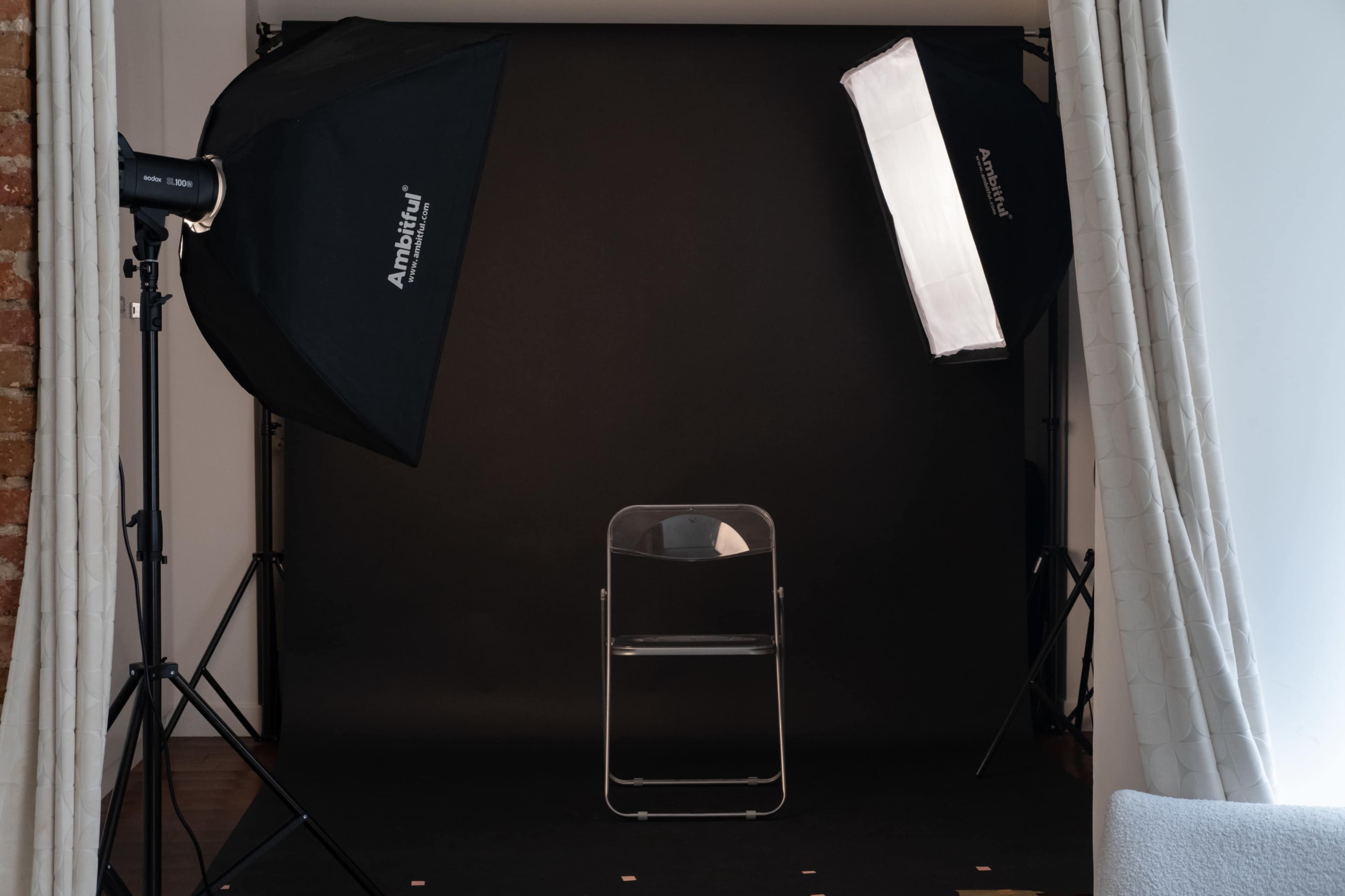 The image shows a photography studio setup featuring a clear chair positioned in front of a black backdrop, with two softbox lights on either side.