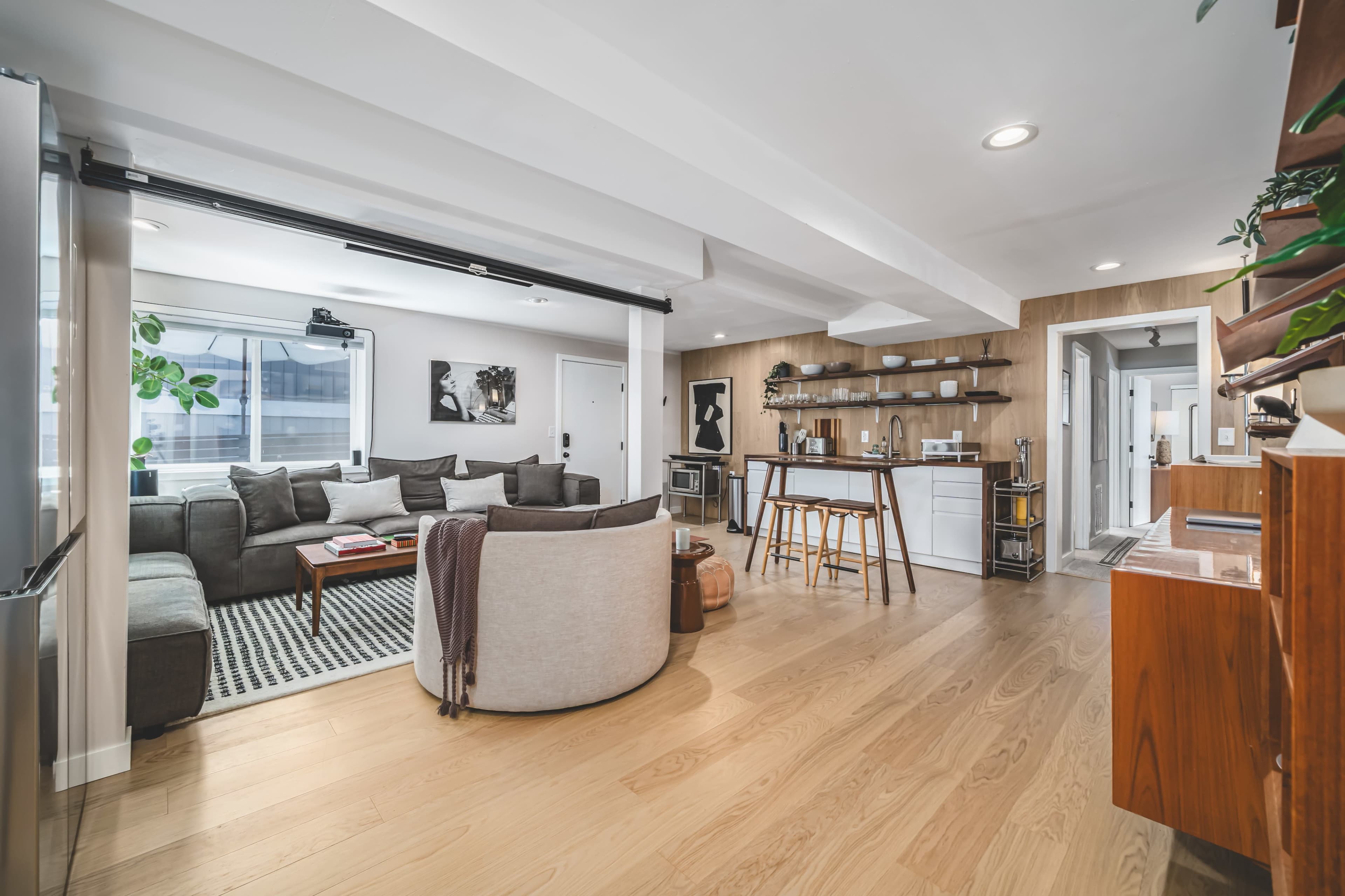 The image shows a modern, open-concept living area featuring a gray sectional sofa, a round chair, a wooden coffee table, and a kitchen area with bar stools.