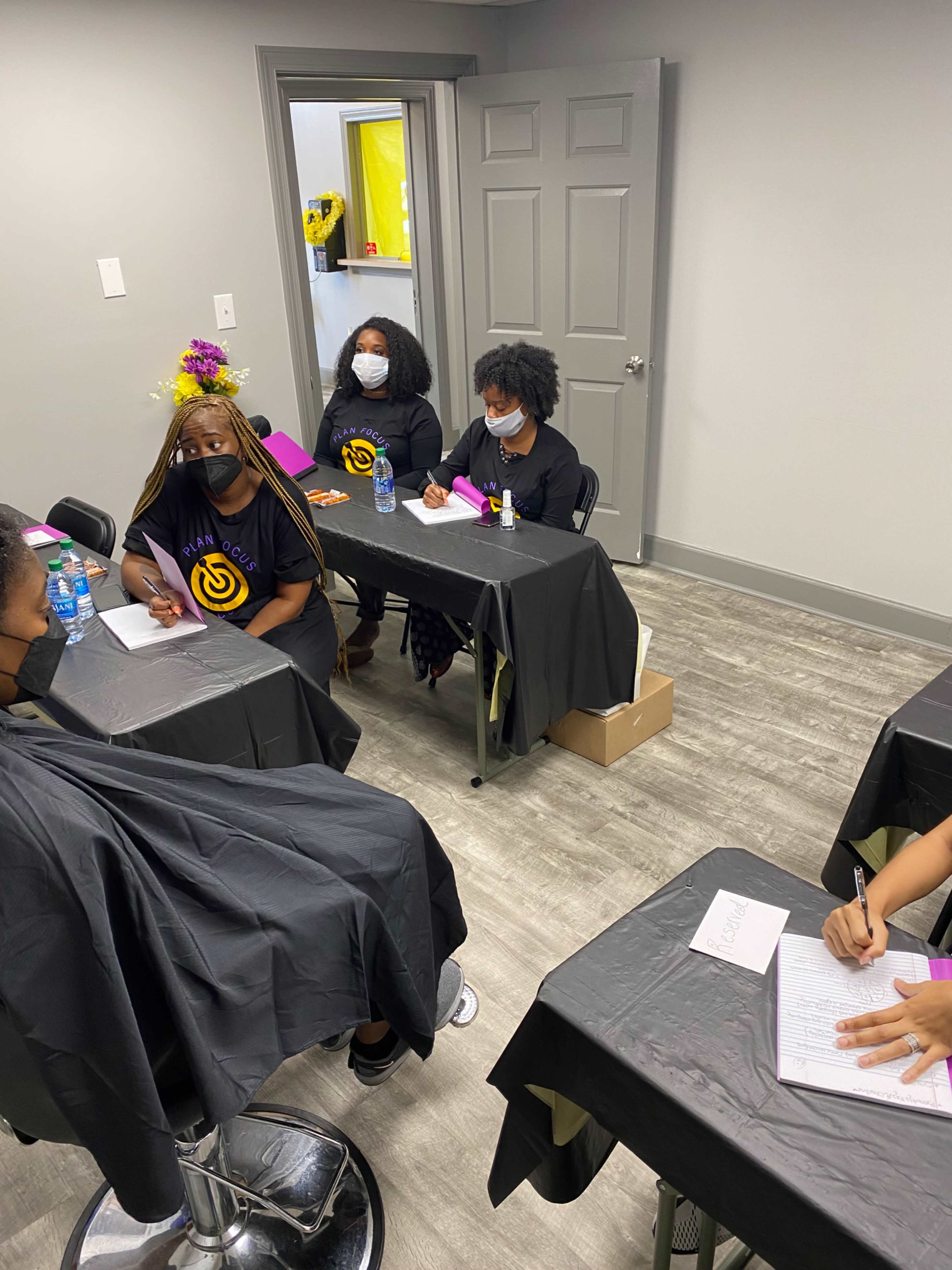 A group of people wearing masks sits at tables in a room with gray walls, engaged in writing and discussion.