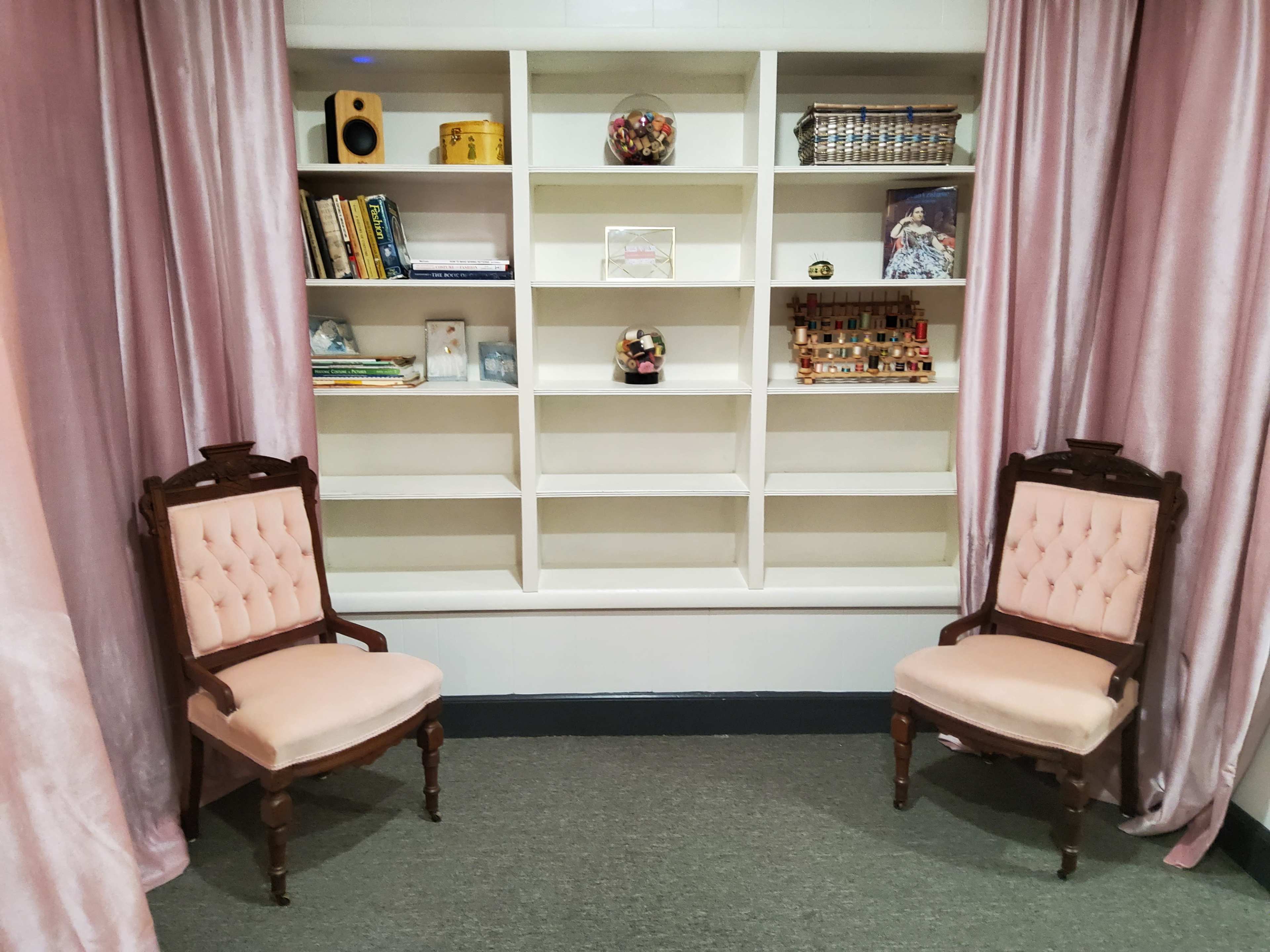 Two upholstered chairs are positioned facing each other in front of a white shelving unit adorned with various items and books, all framed by soft, pink curtains.