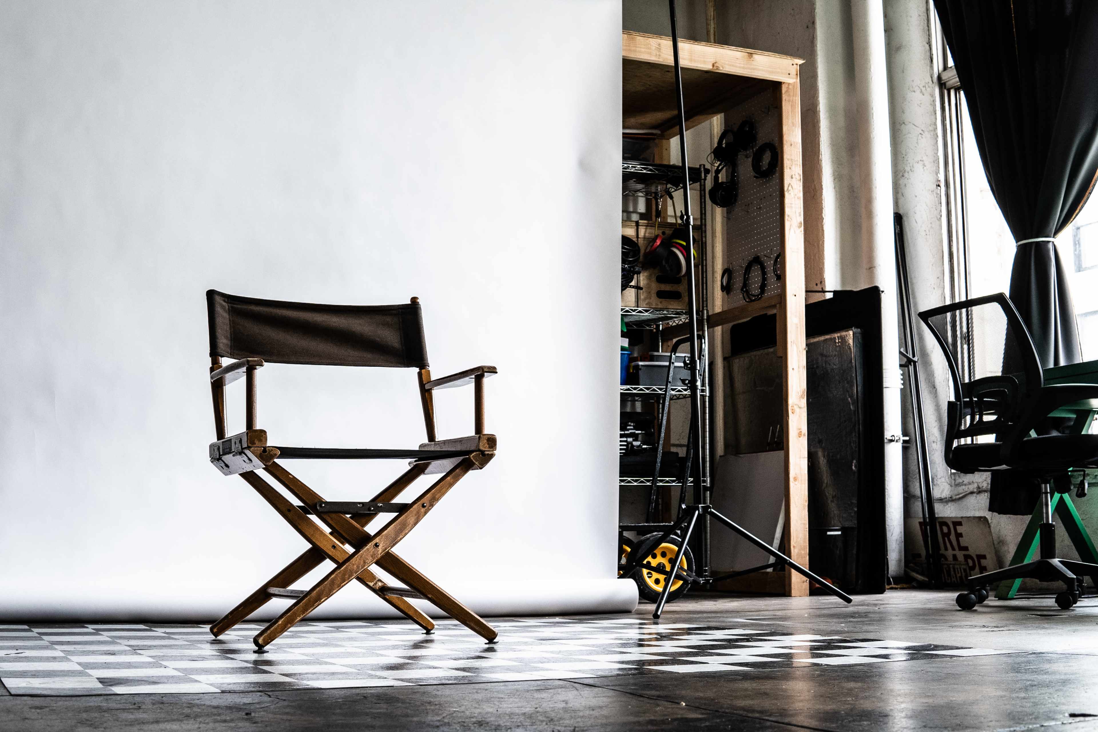 A wooden director's chair is positioned in front of a large white backdrop in a studio with shelves and equipment in the background.