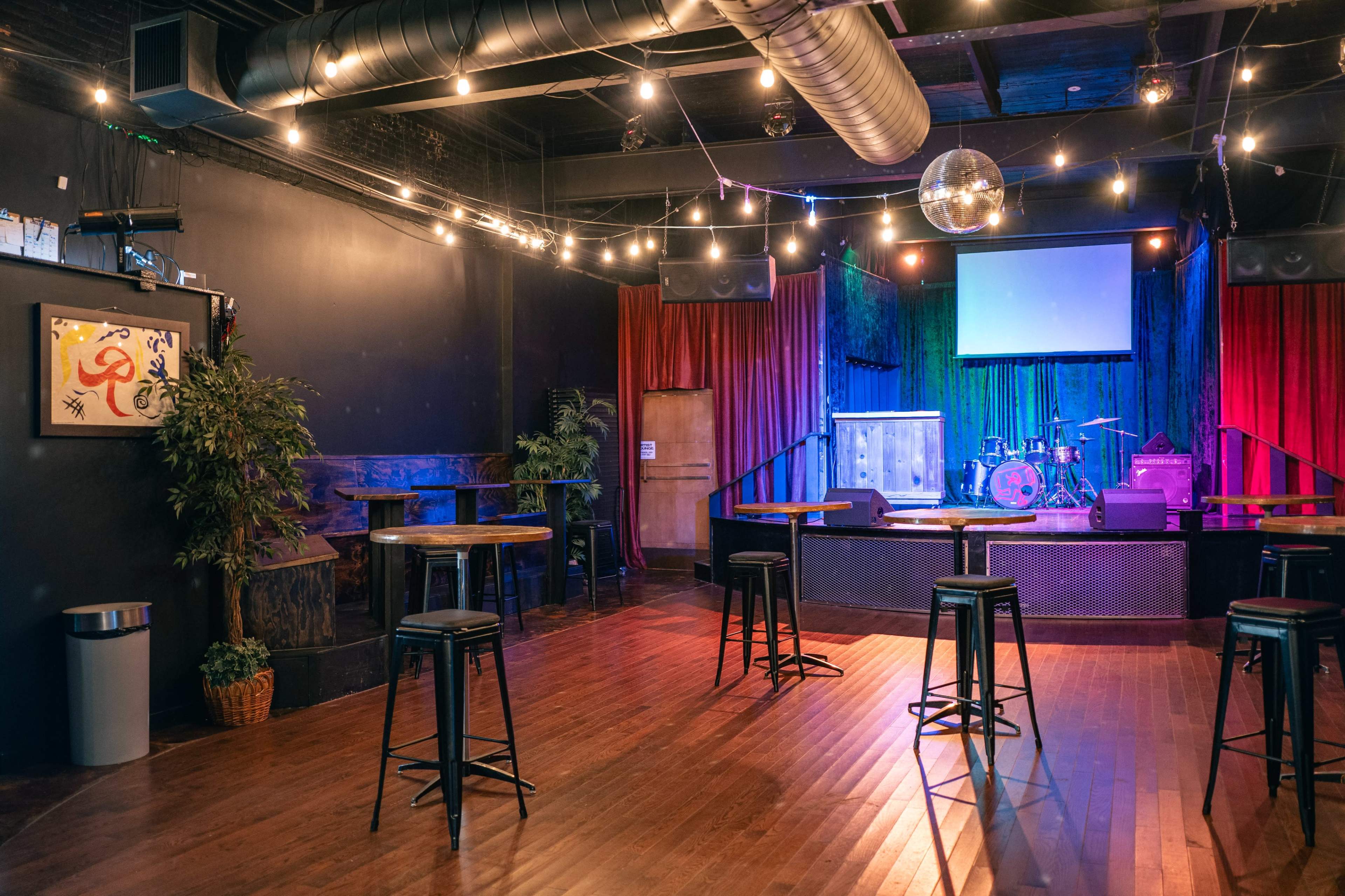 The image shows an empty music venue featuring a stage with instruments, a projector screen, and scattered high-top tables under warm, ambient lighting.