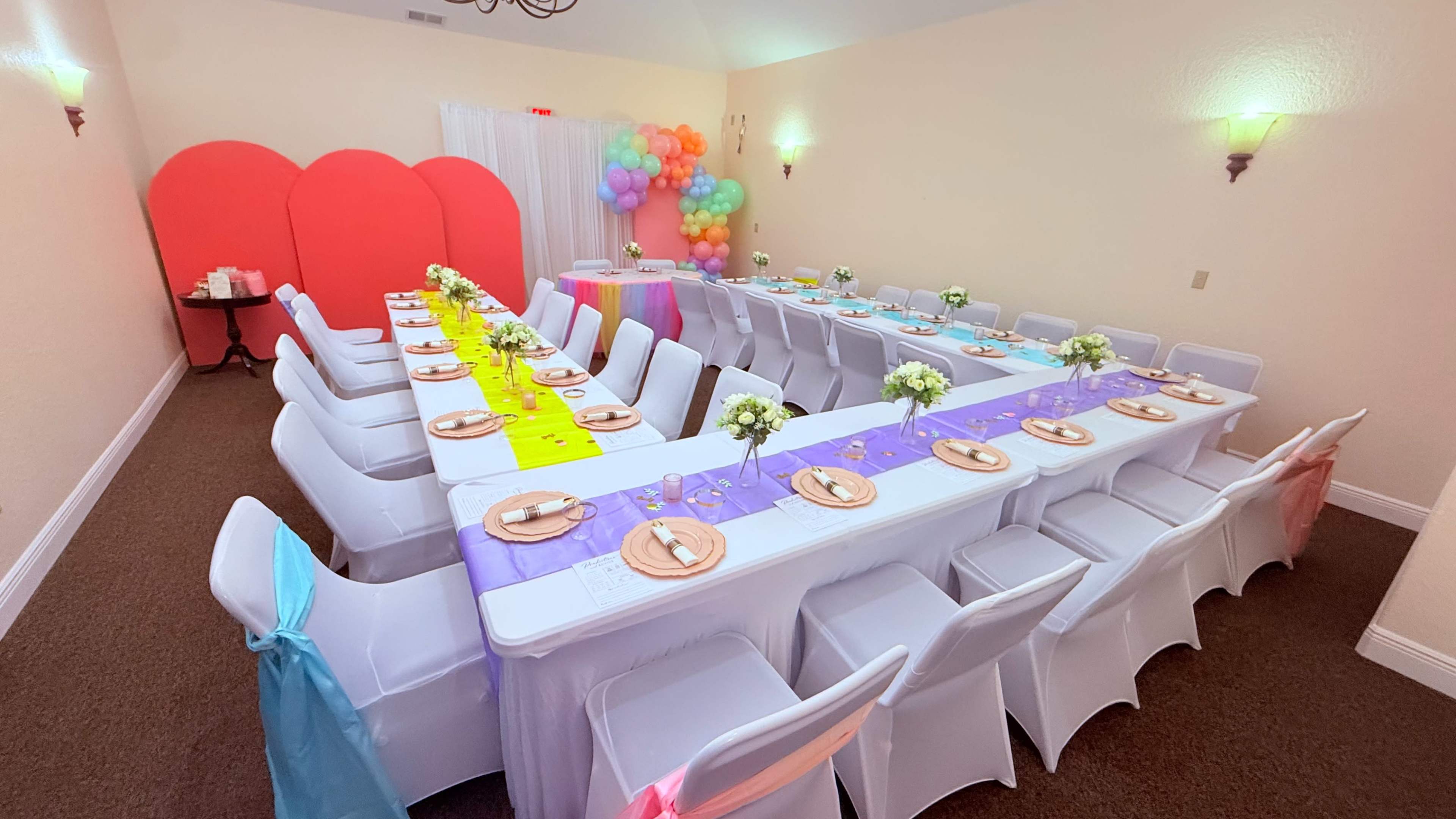 A brightly decorated banquet room features a long table set with white tablecloths, colored runners, and floral centerpieces, surrounded by chairs adorned with sashes.