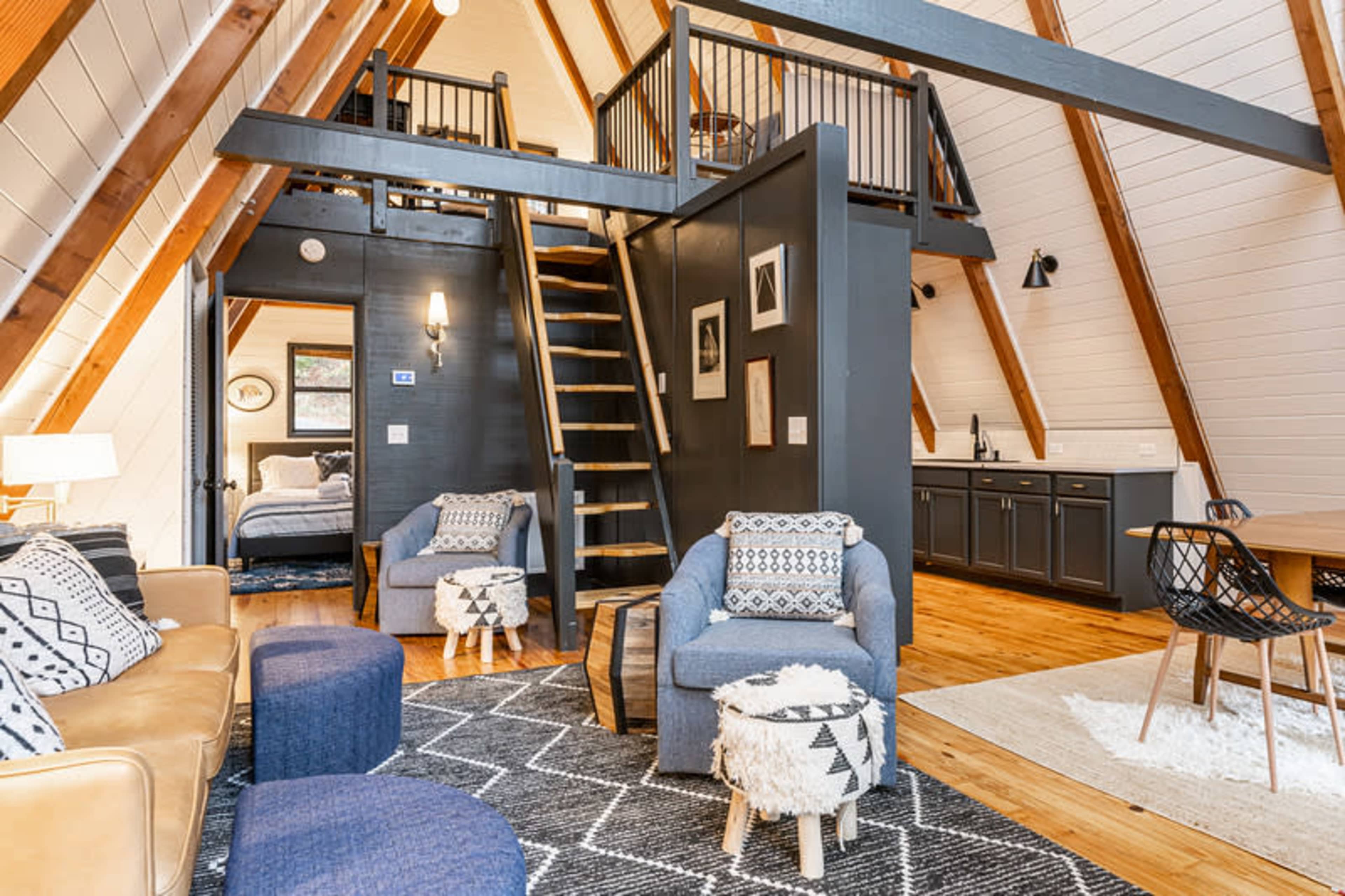 The image shows a cozy, vertical living space with a wooden staircase leading to a loft area, featuring a seating arrangement with two chairs, a sofa, and a kitchen area in the background.
