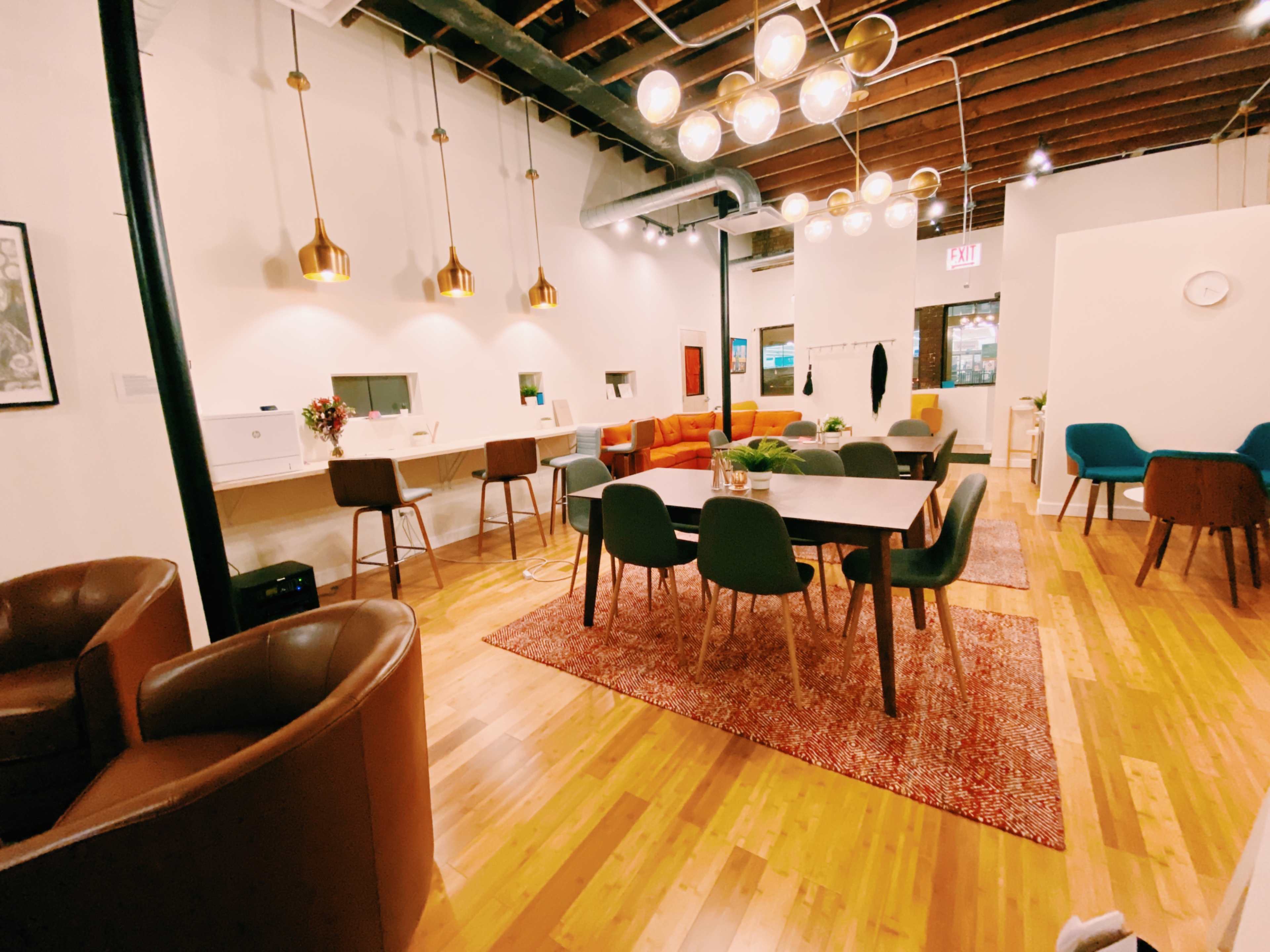 The image shows a modern, open-concept interior space featuring a dining area with a large table, a lounge area with colorful seating, and decorative light fixtures hanging from the ceiling.