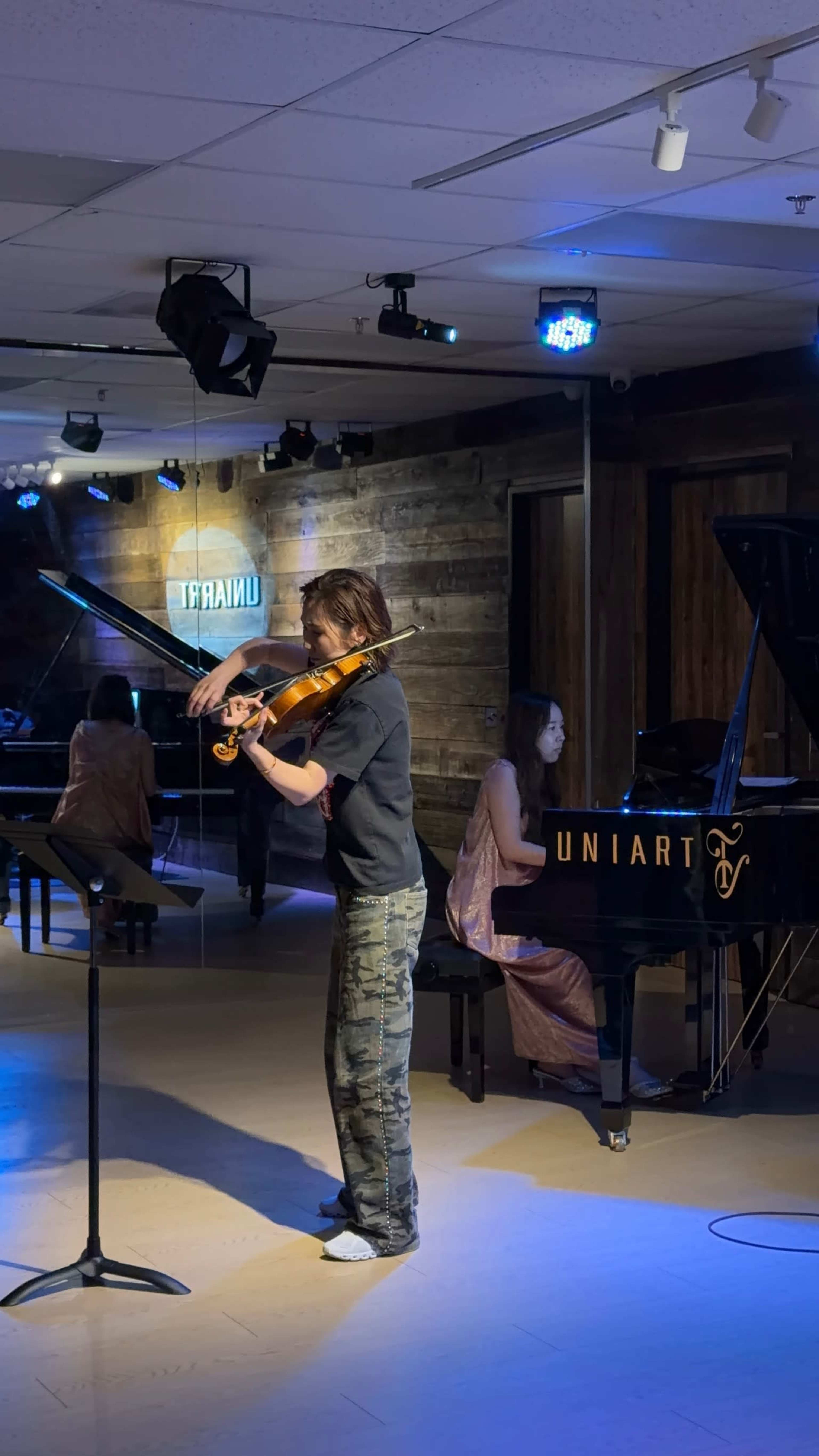 A young violinist performs on stage while a pianist accompanies them in a dimly lit venue.