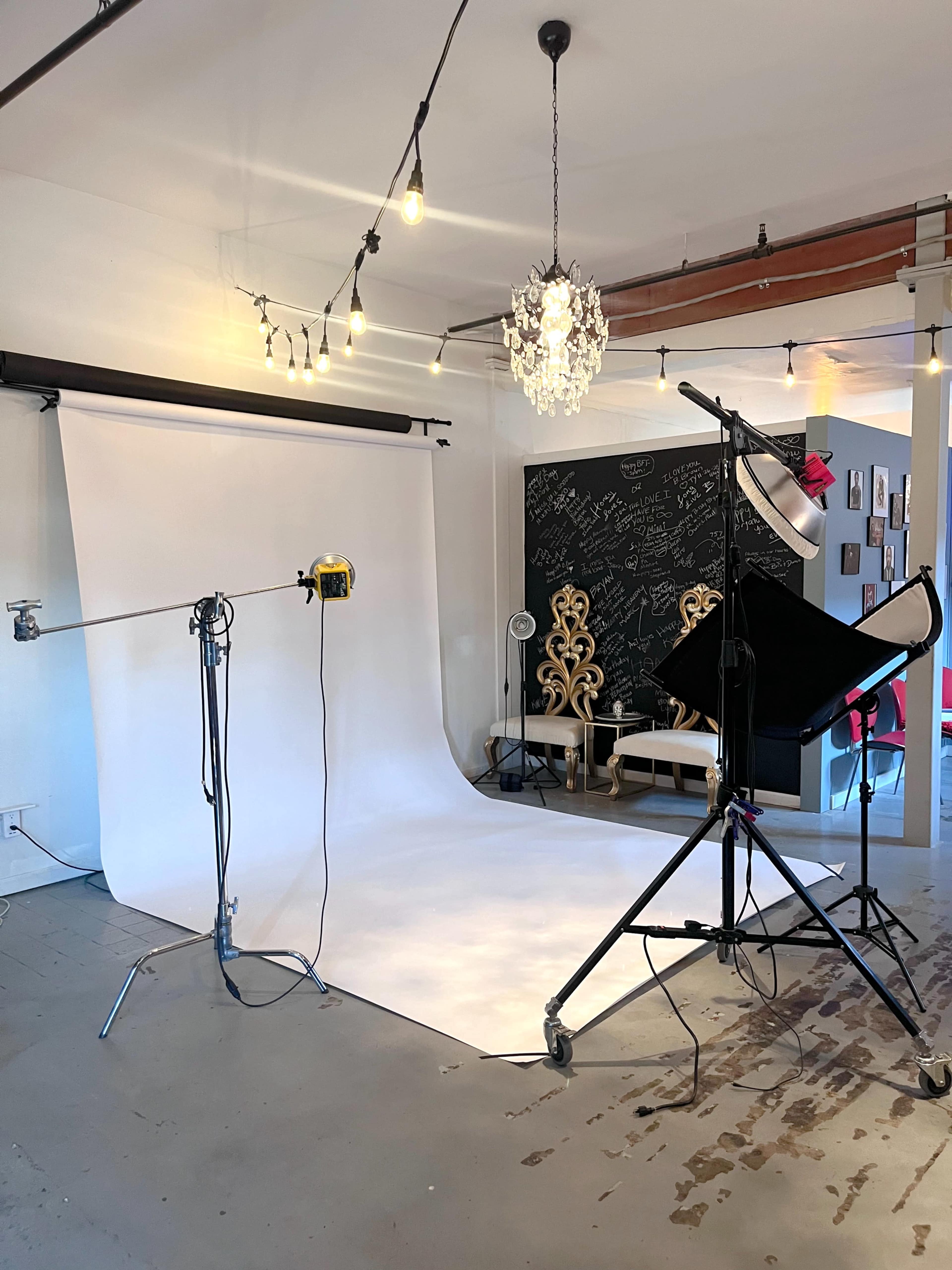 The image shows a photography studio with a white backdrop, lighting equipment, and a decorative black wall with various framed pictures.