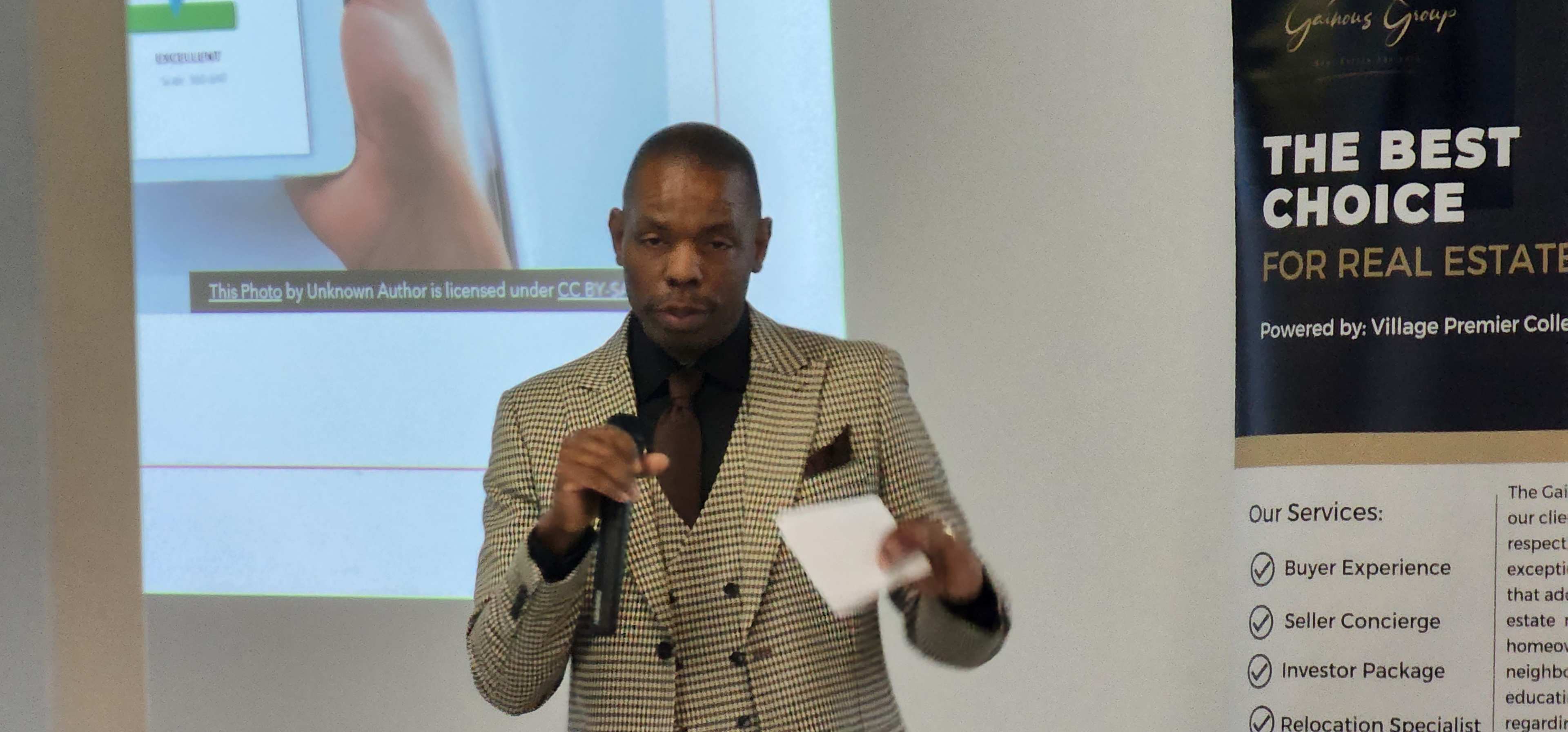 A man in a patterned suit speaks into a microphone during a presentation at a real estate event.