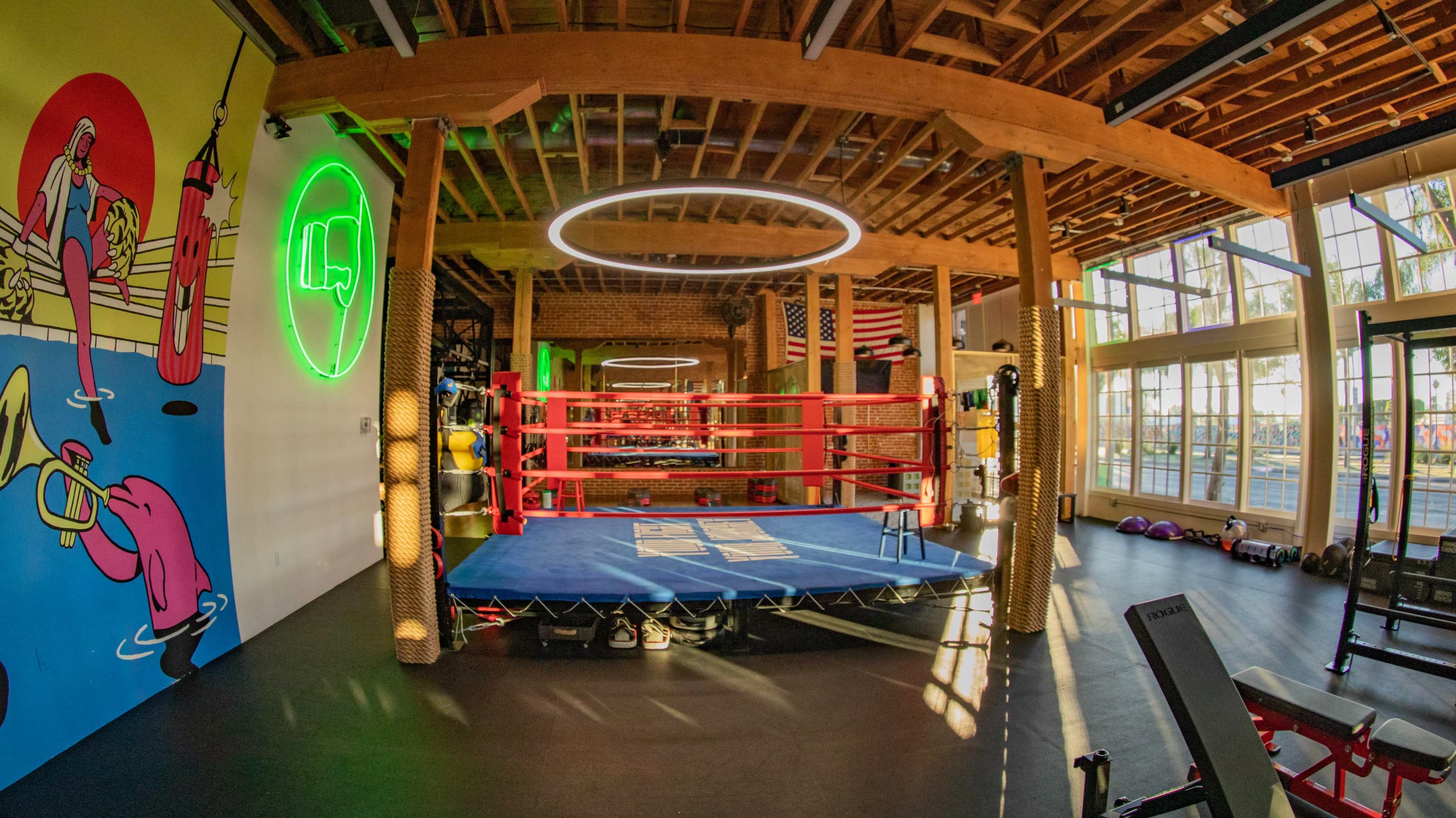 A boxing gym features a central ring surrounded by vibrant wall art, seating, and fitness equipment.