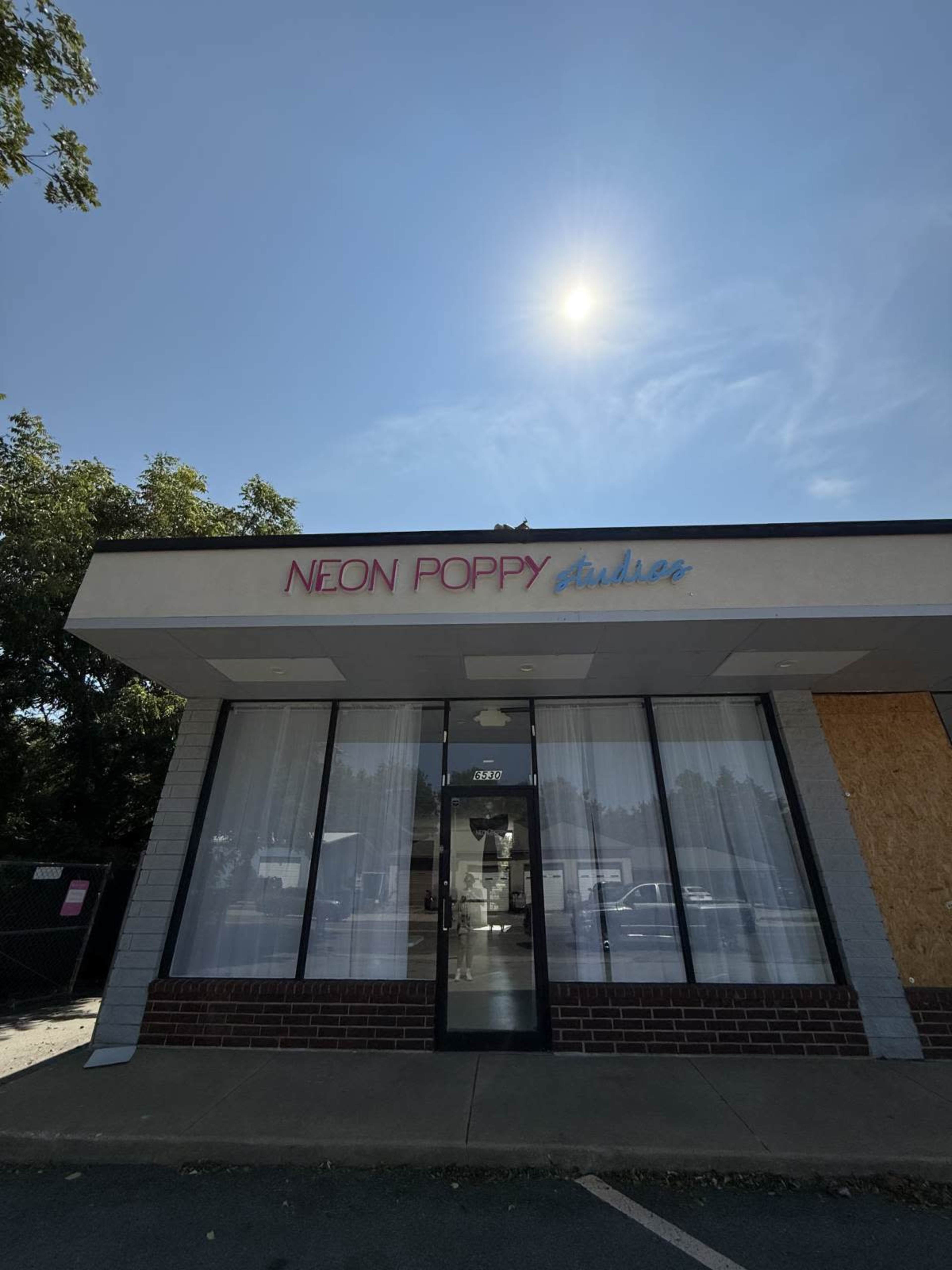 The image shows a storefront with the sign "NEON POPPY studios" on a clear day, featuring large windows and a partially boarded-up area.