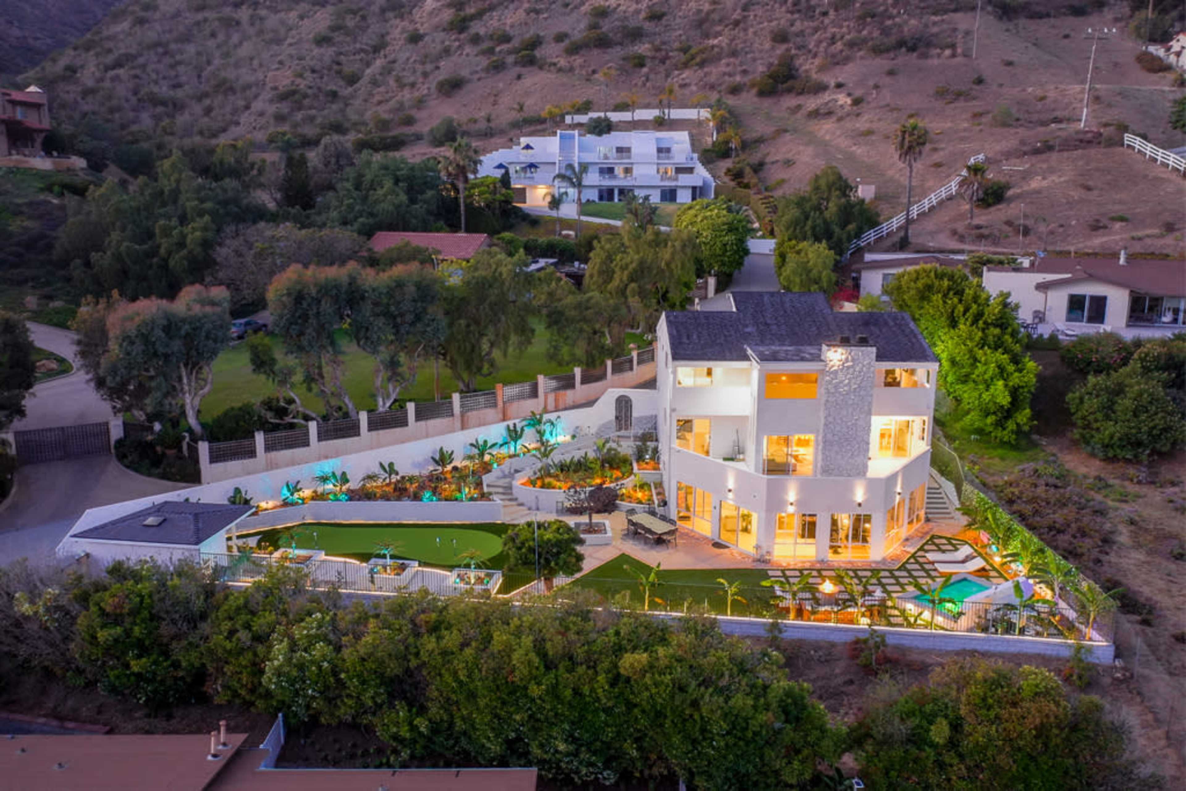 The image shows a modern house with large windows, set on a landscaped property featuring a pool and surrounded by hilly terrain.