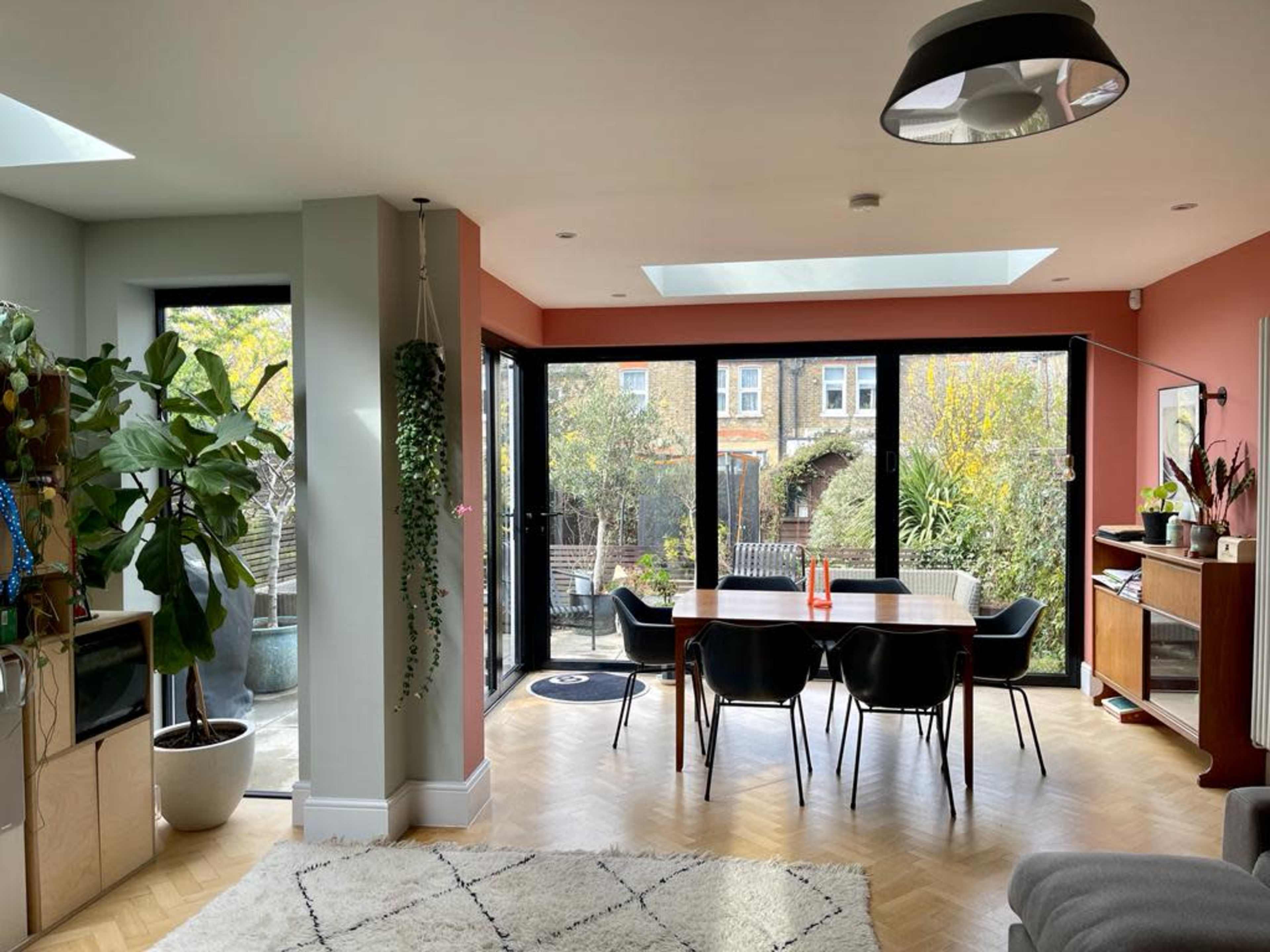 The image shows a modern dining area with large glass doors opening to a garden, featuring a wooden table surrounded by black chairs and potted plants.