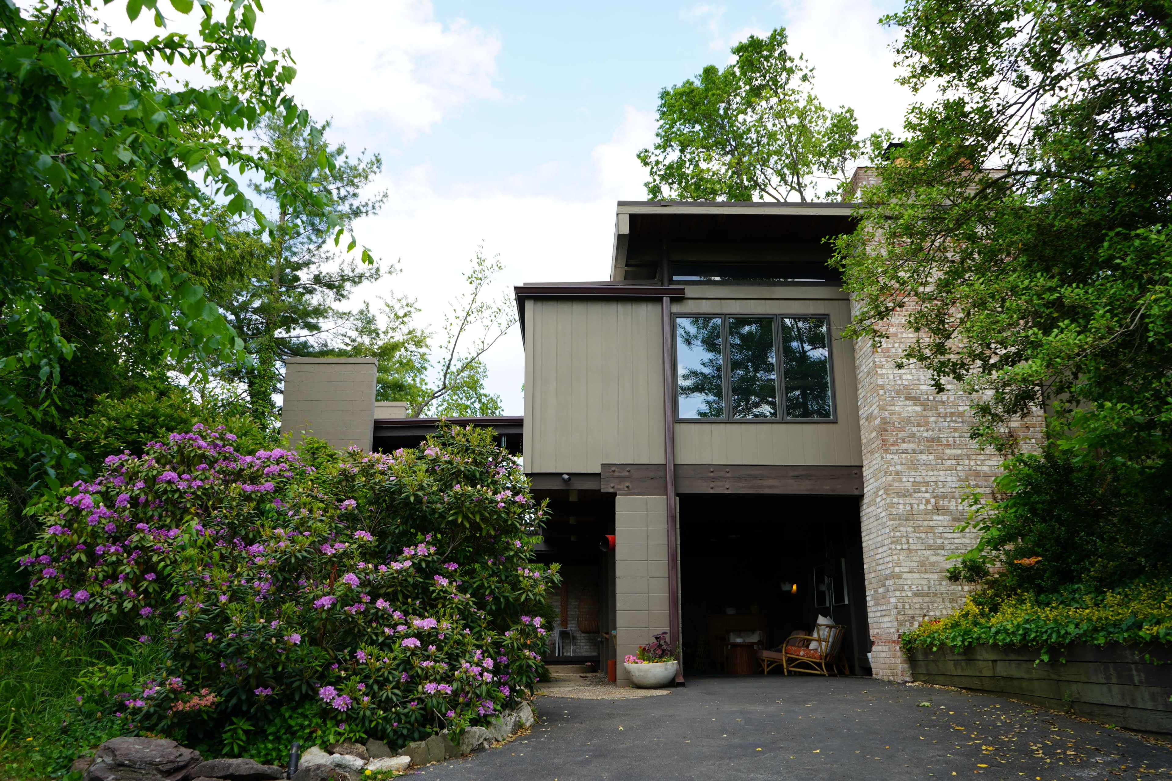 A modern two-story house with large windows and a stone facade is surrounded by a garden of blooming flowers.