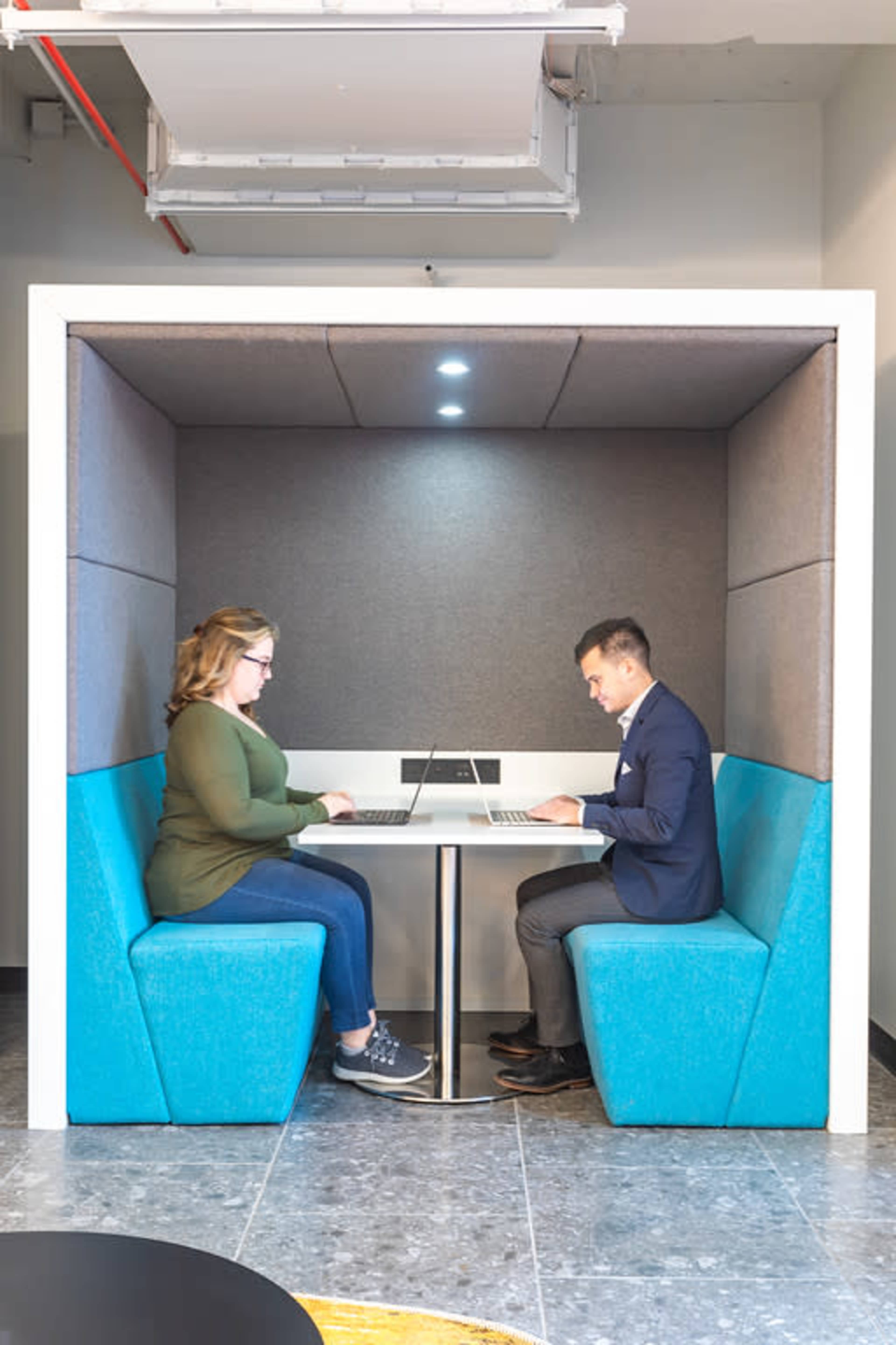 A woman and a man are sitting across from each other at a small table in a modern workspace pod, each using a laptop.