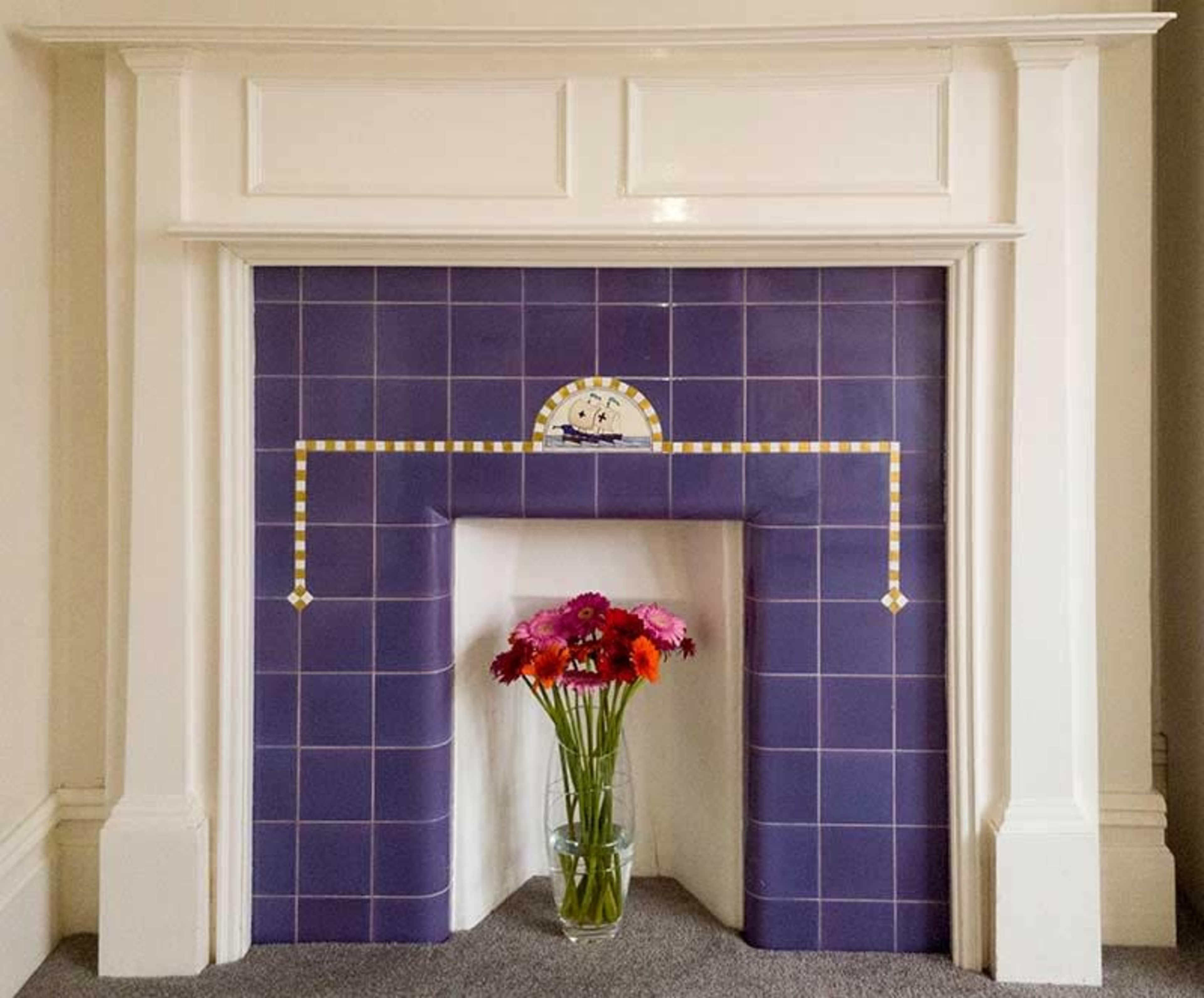A decorated fireplace with a purple tile surround and a vase of colorful flowers in front of it.