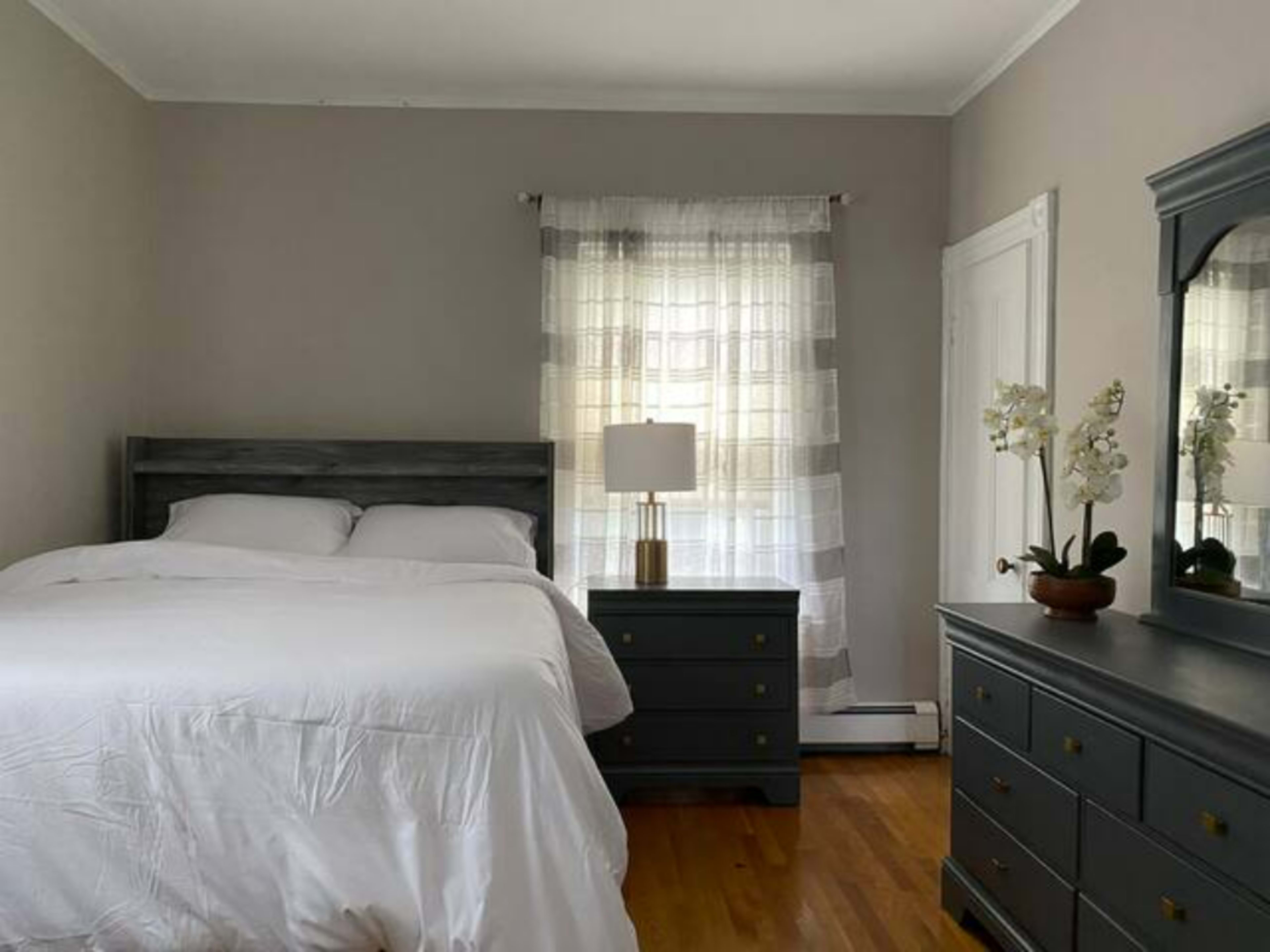 A well-organized bedroom features a bed, a nightstand with a lamp, and a dresser, all set against light gray walls and natural light filtering through a curtain.