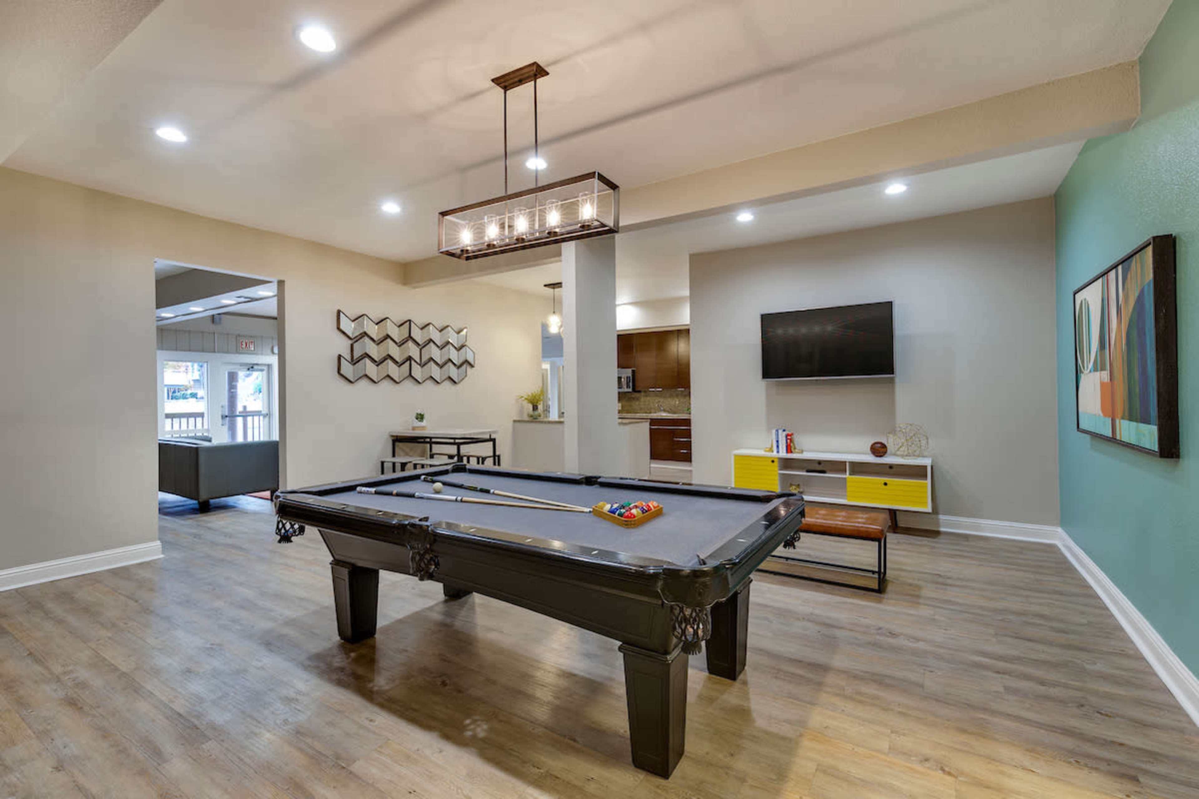 A modern game room featuring a billiards table, a flat-screen TV, and various seating arrangements with wood flooring and pendant lighting.