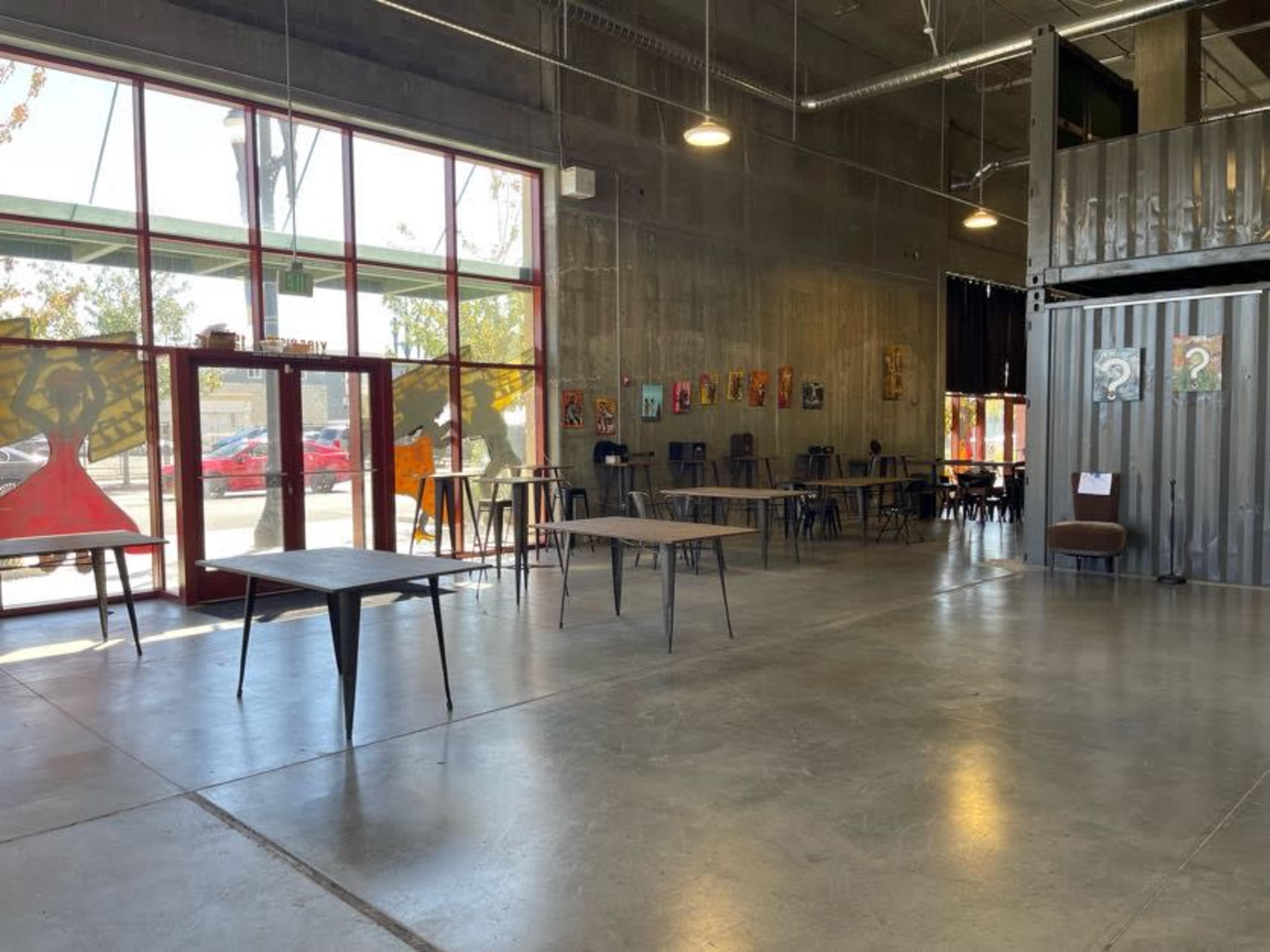 An indoor space with concrete floors, large windows, and several tables arranged throughout the area, along with artwork hung on the walls.