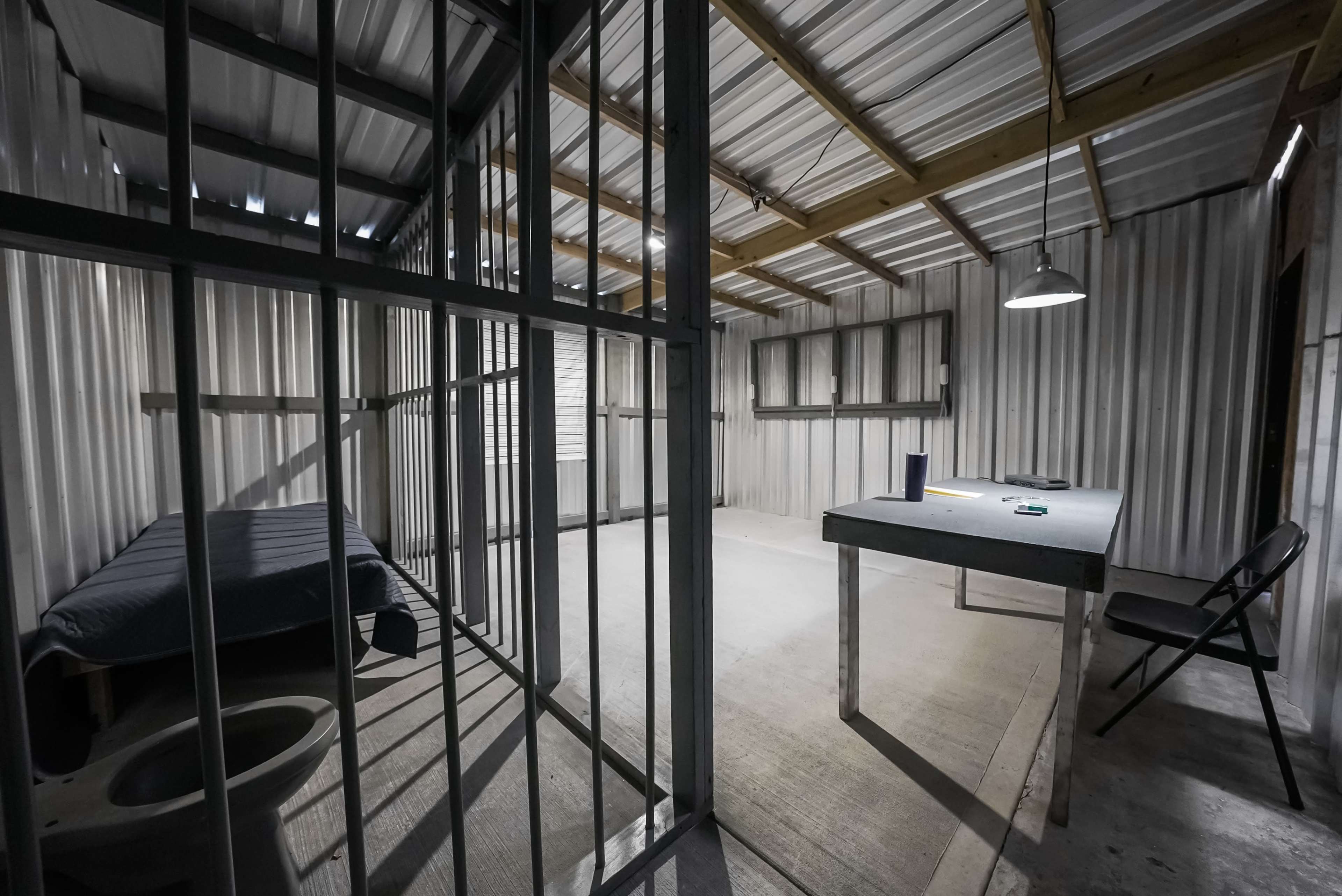 The image shows a dimly lit prison cell with a bed on one side and a table with a chair on the other, both enclosed by metal bars.