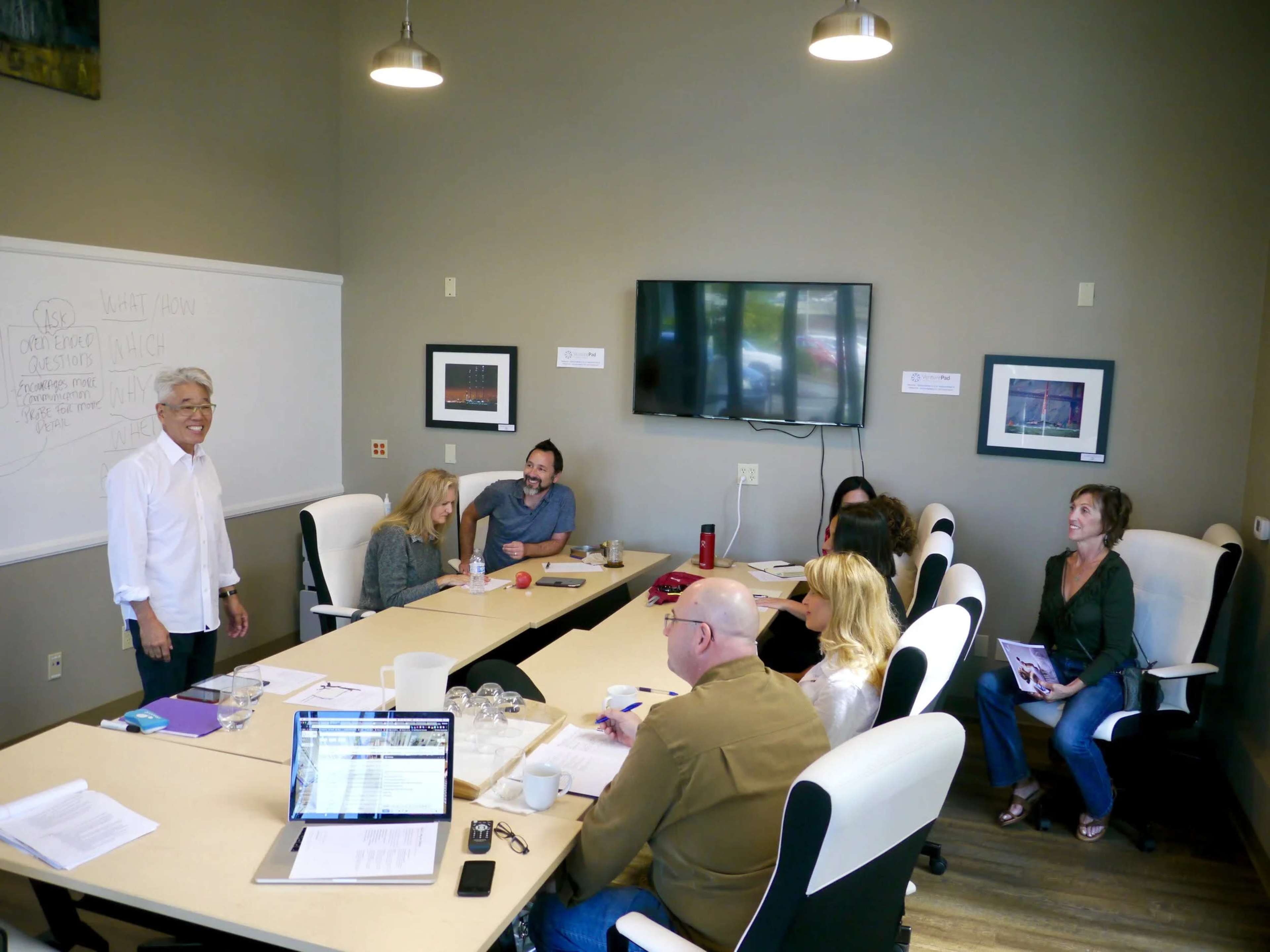 A group of seven people is seated around a conference table in a meeting room, while one person stands at the front discussing a chart on a whiteboard.