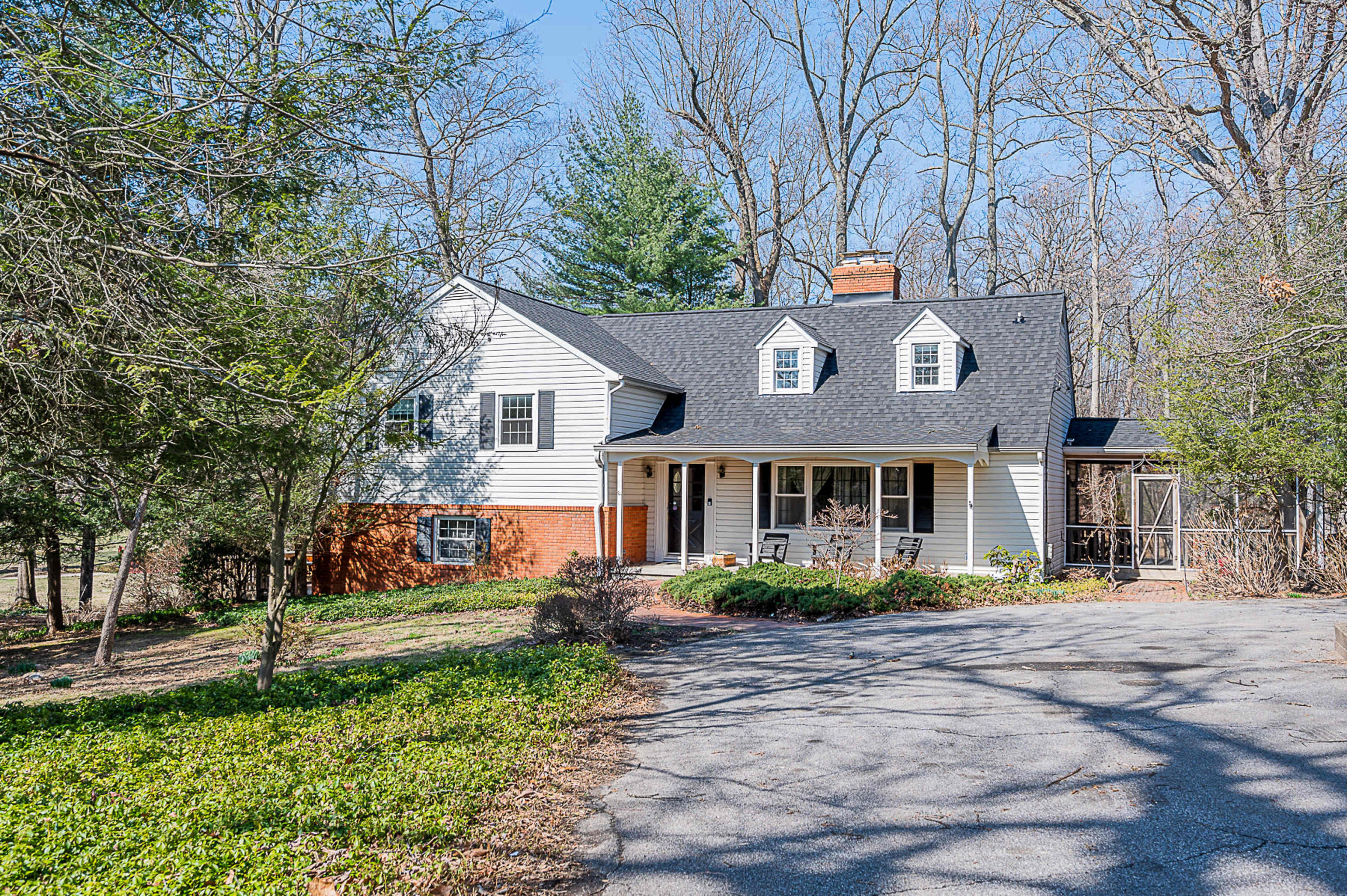 A two-story, white-clapboard farmhouse with a dark roof and a brick foundation is situated on a wooded lot.