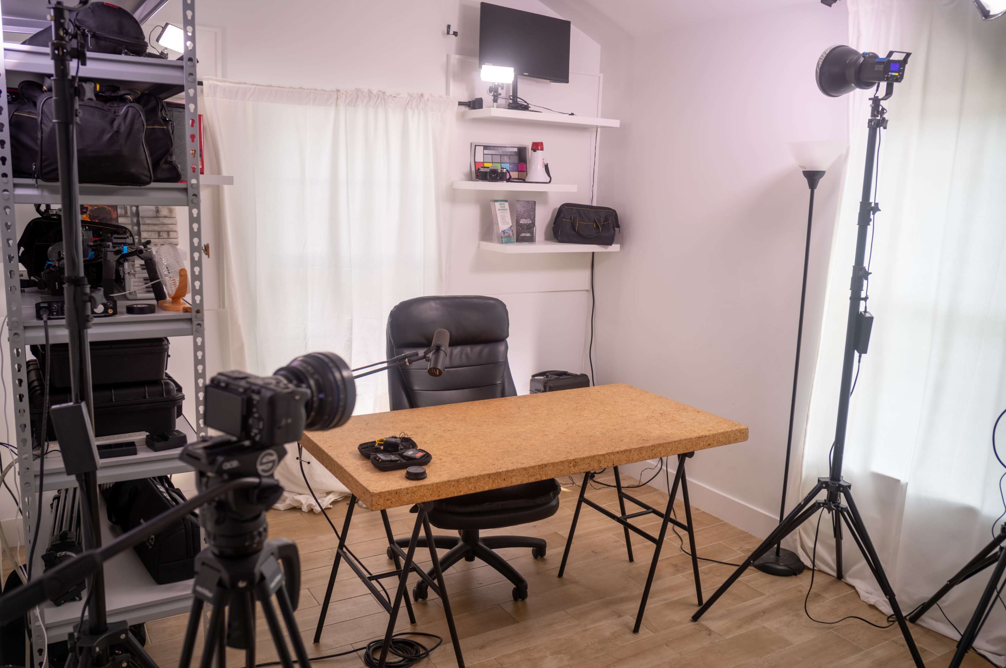 A well-lit room featuring a desk and a chair, with various camera equipment and lighting setup on either side.