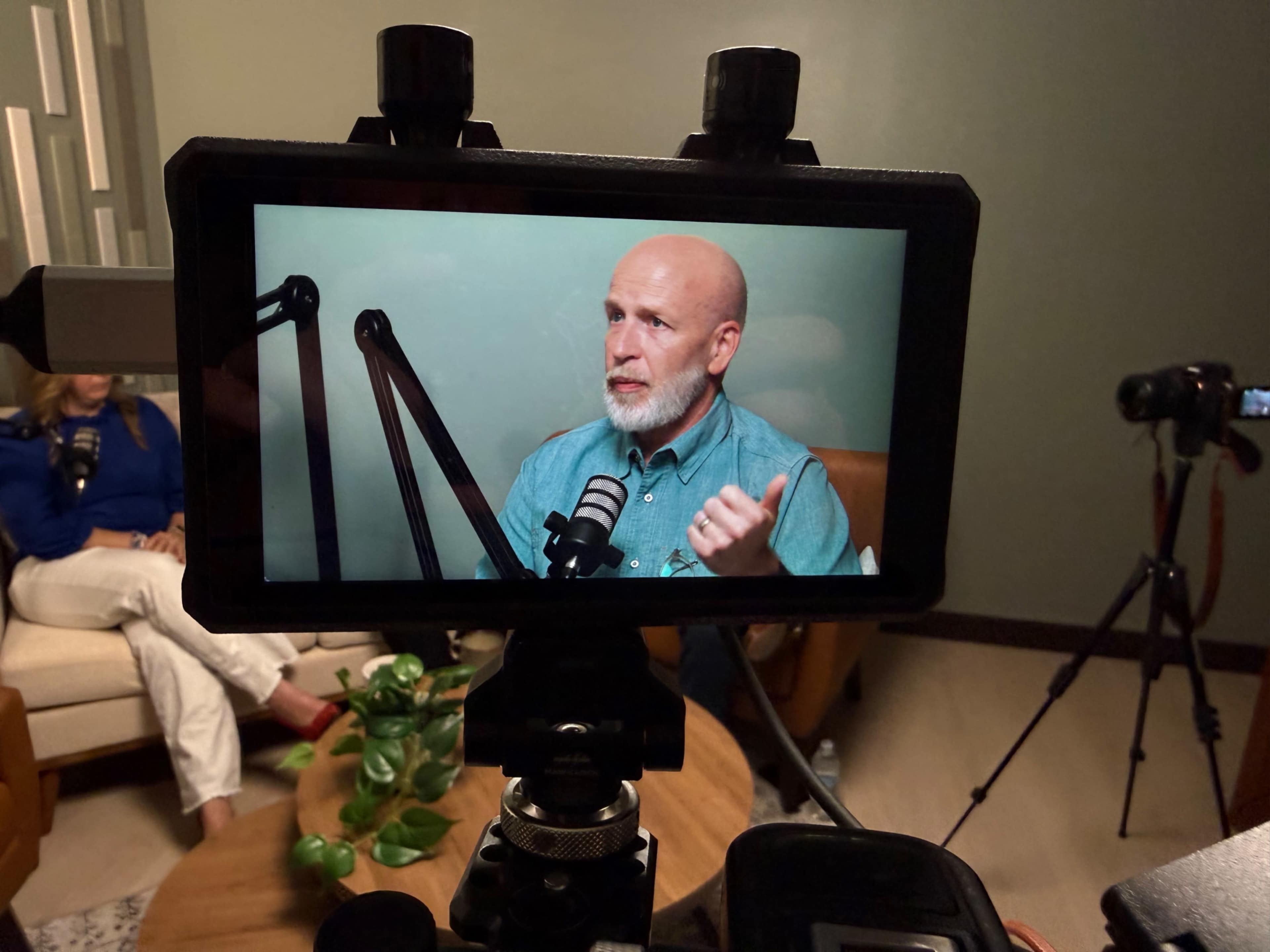 A man with a beard speaks into a microphone while seated in a studio, with a camera focused on him.