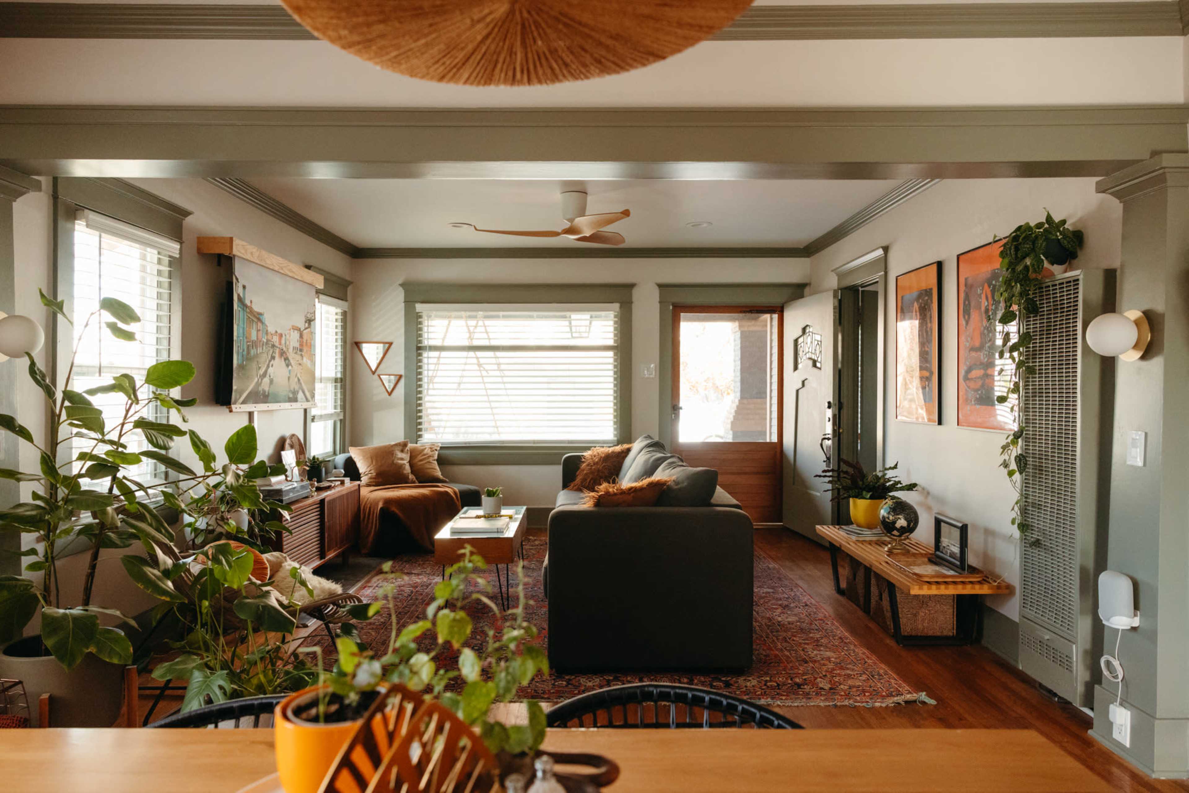 A cozy living room with plants, a sofa, a coffee table, and large windows providing natural light.