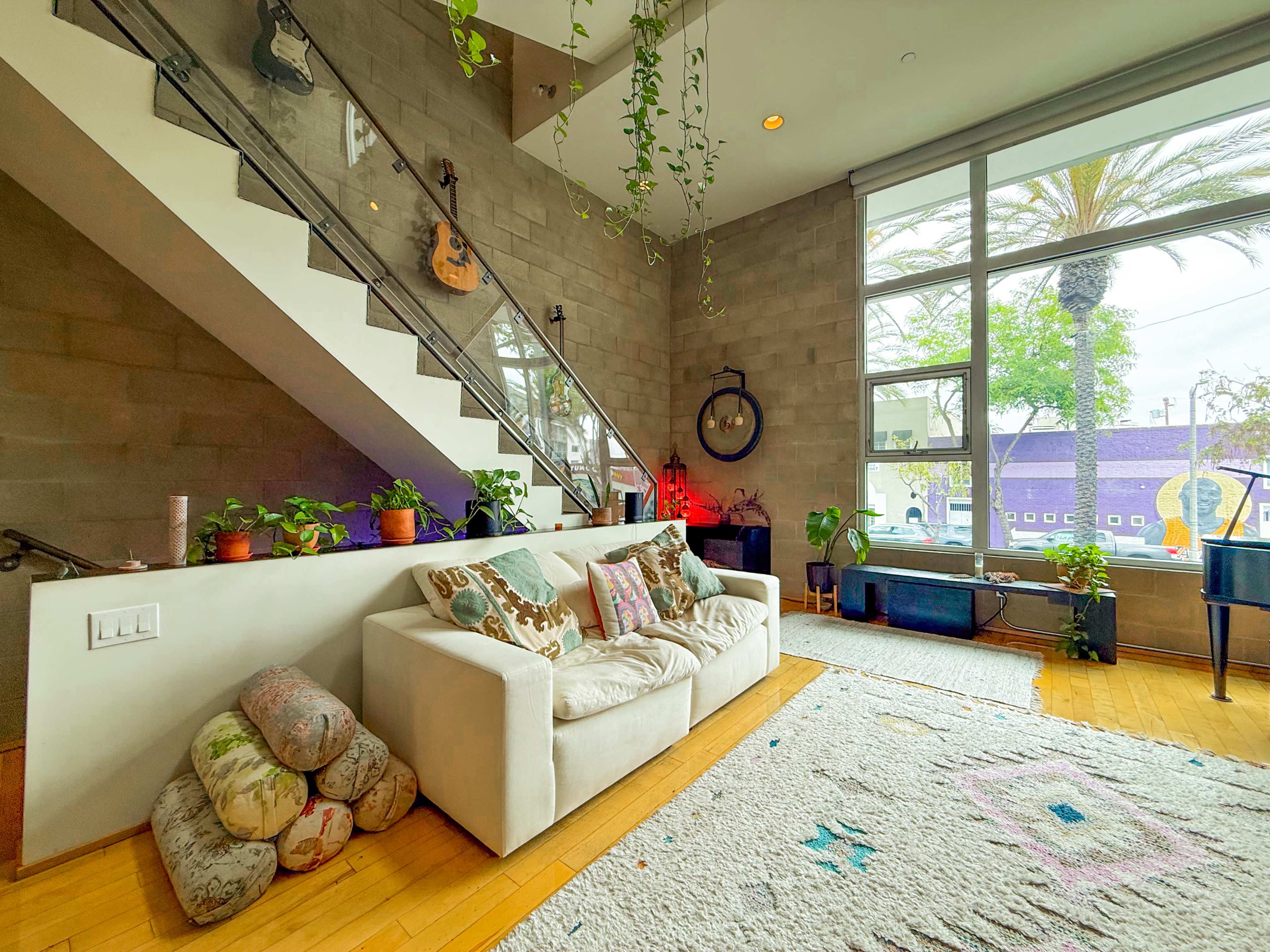 The image shows a modern living space with a white couch, potted plants, and a large window featuring palm trees outside, complemented by a staircase leading to an upper level.