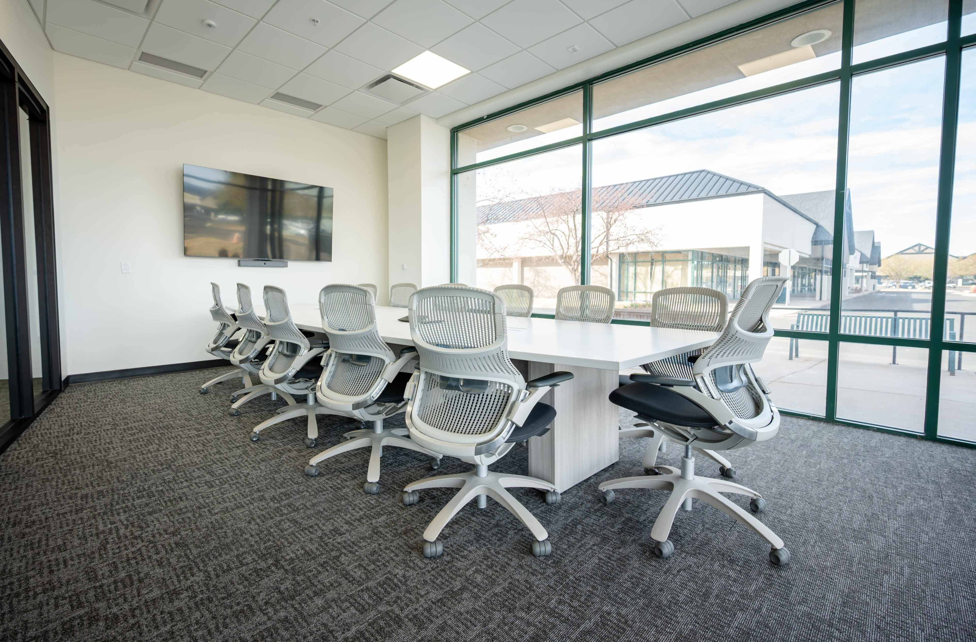 A modern conference room features a long table surrounded by ergonomic chairs, with large windows providing a view of the outside.