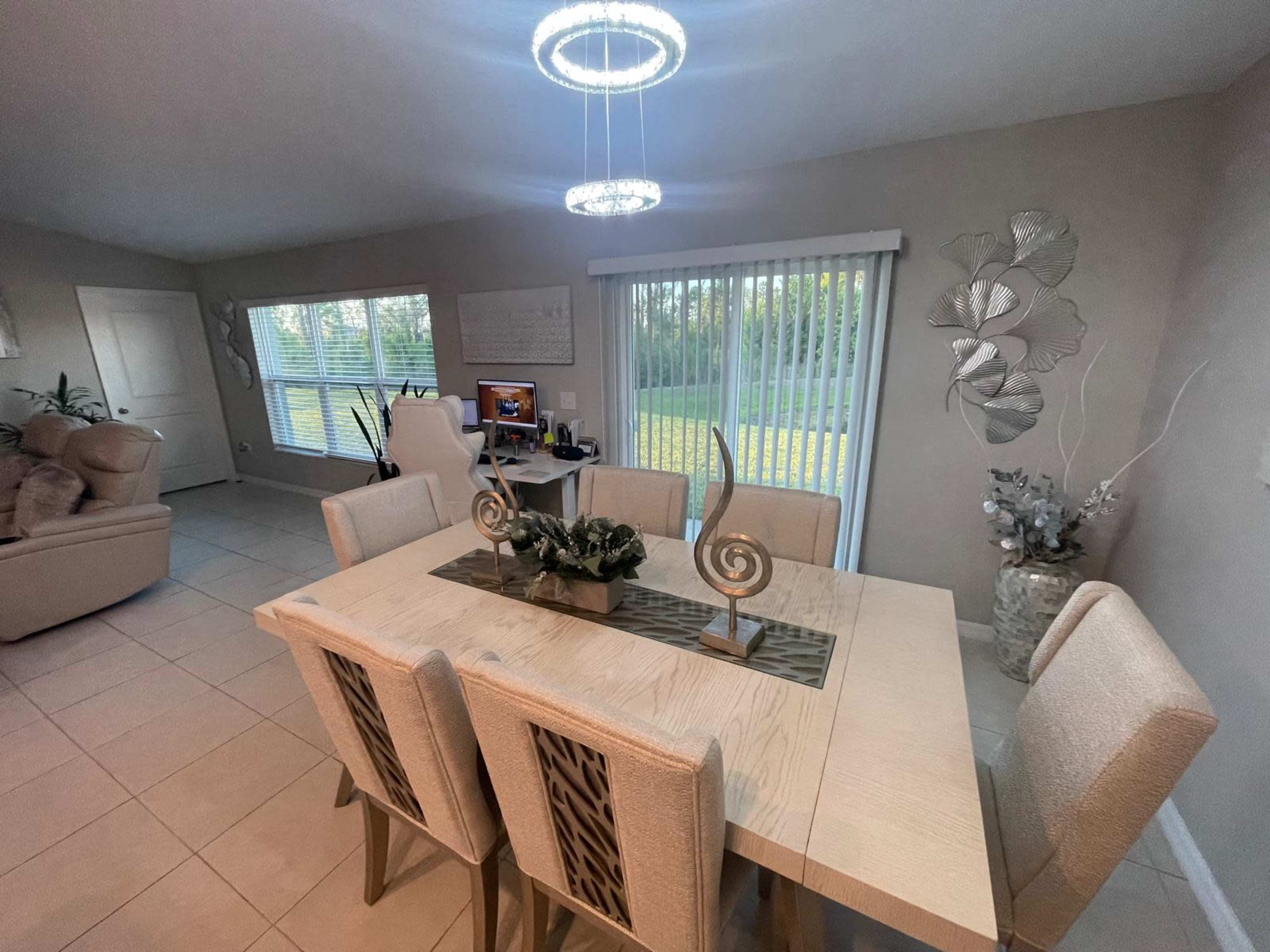 The image shows a modern dining room with a light-colored table and matching upholstered chairs, a decorative centerpiece, and large windows allowing natural light to enter the space.