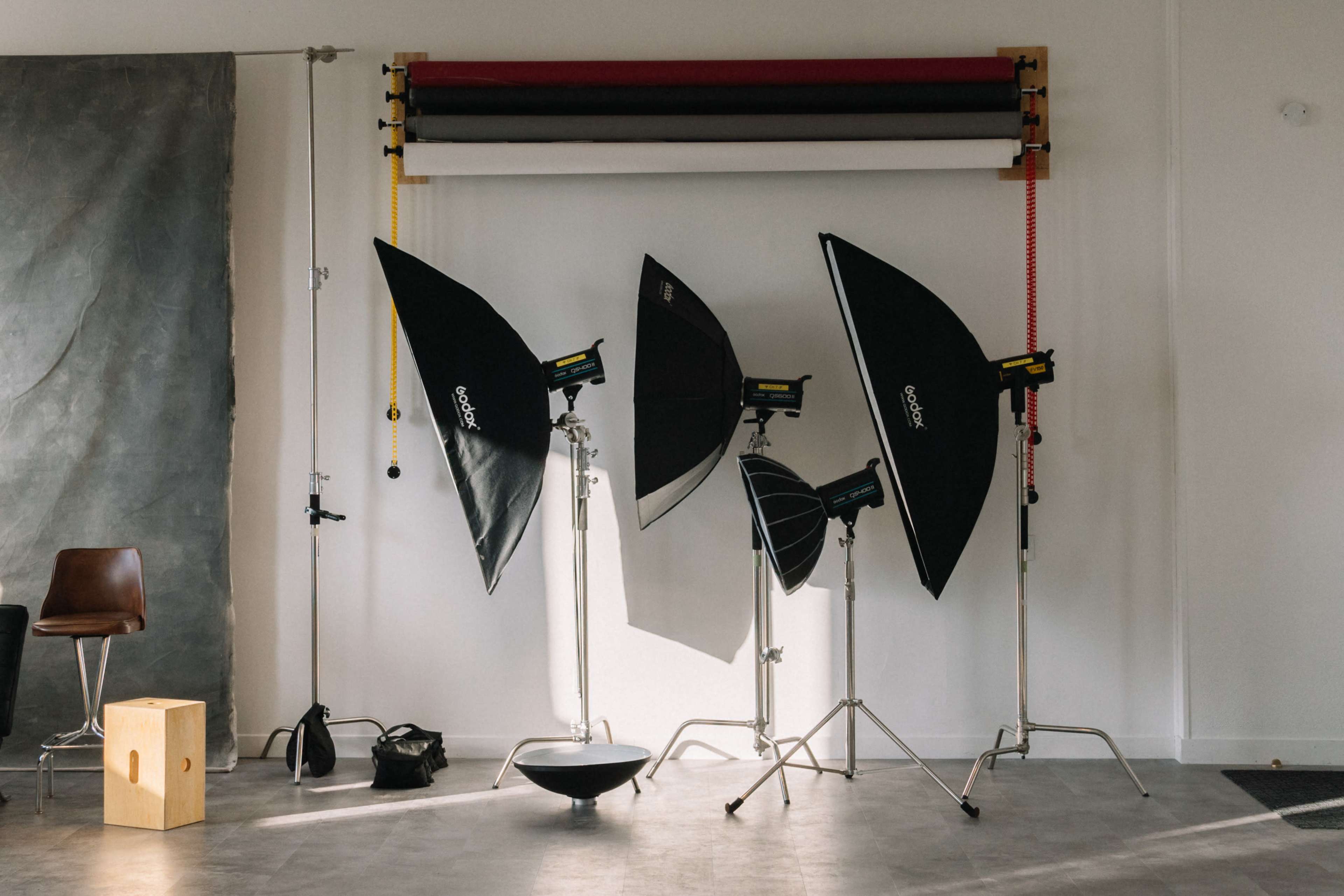 A photography studio setup with several softbox lights, a backdrop roller, and various equipment arranged against a wall.