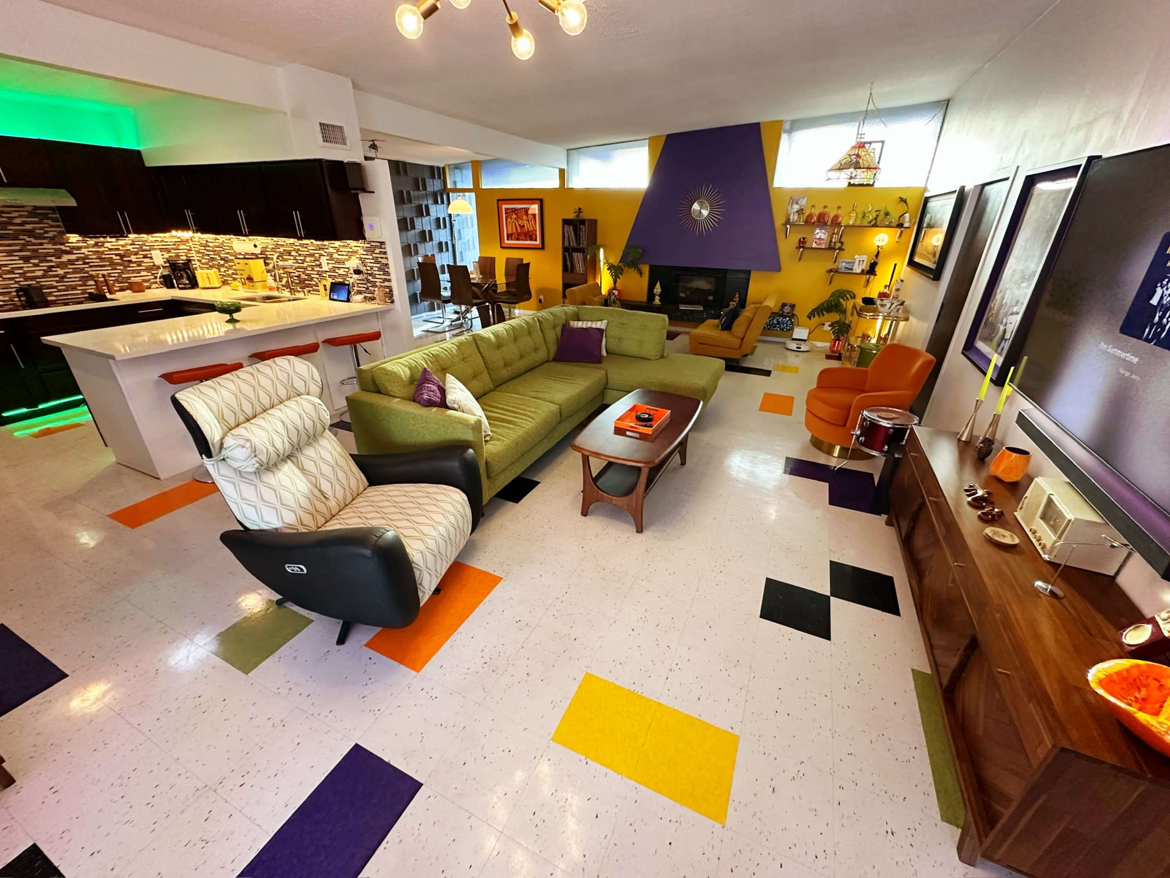 The image shows a brightly colored modern living space featuring an open layout with a green sectional sofa, a wooden coffee table, and various decorative elements on the walls and shelves.
