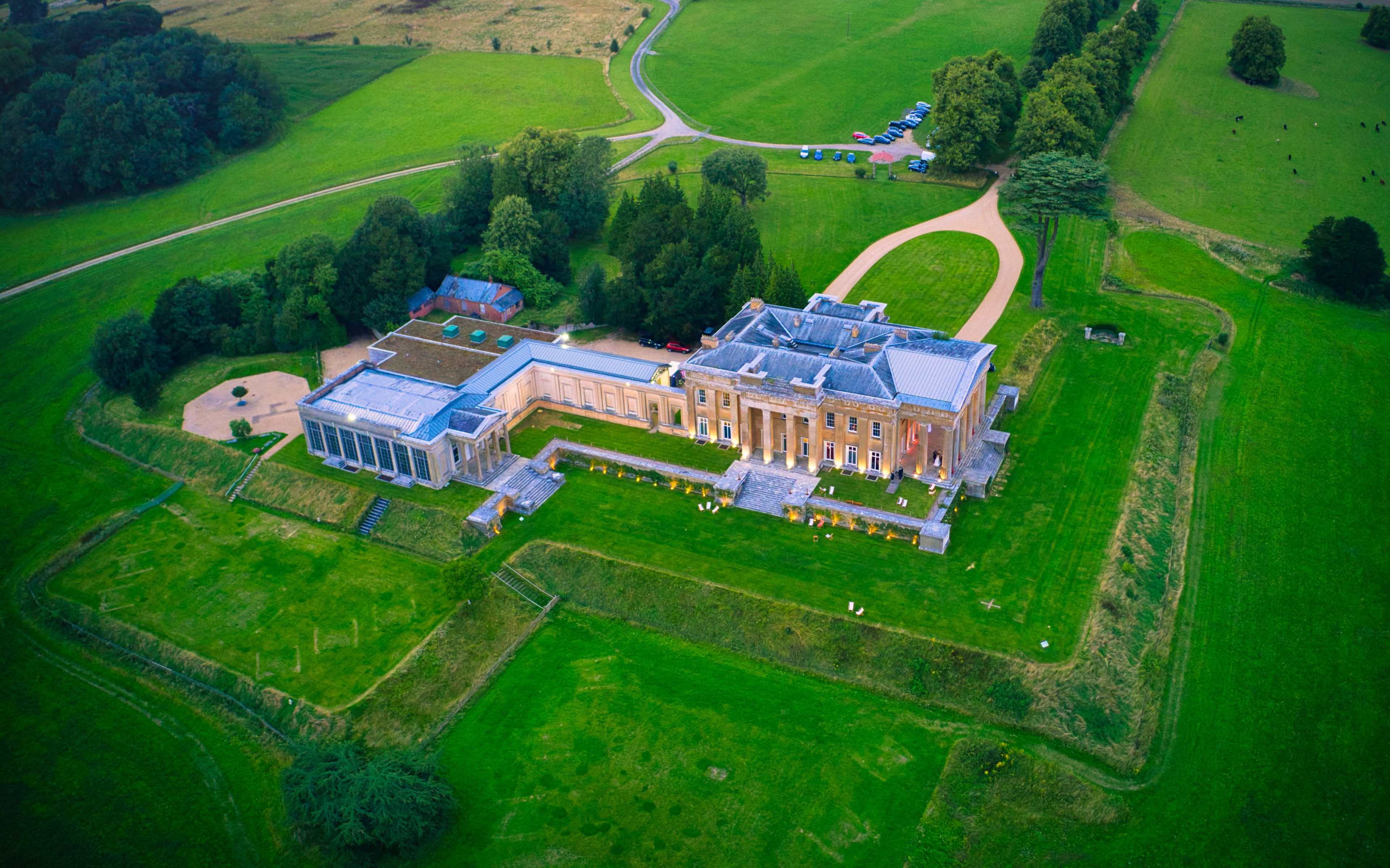 The image shows a large mansion situated on a sprawling green landscape with a winding road leading to it, surrounded by trees and open fields.